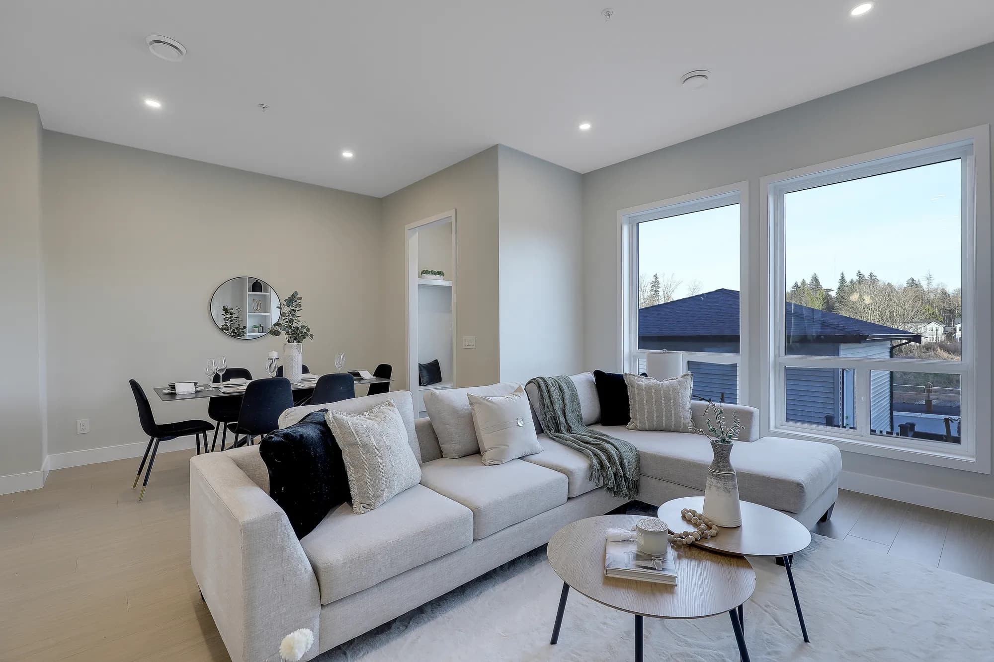 Latimer Landing – A modern open-concept living and dining area featuring a light grey sectional sofa with throw pillows, round wooden coffee tables, and light hardwood floors. Large windows bring in natural daylight while showcasing views of neighboring rooftops and trees, adjacent to a contemporary dining table set for four.