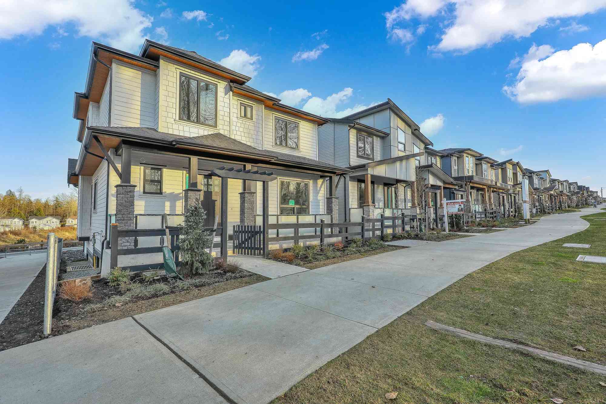 Latimer Landing – A row of modern two-story townhomes features light-toned siding, dark trim, and stone-accented front porches along a paved pedestrian sidewalk. The daytime shot captures neat landscaping, a curving walkway, and clear blue skies with scattered white clouds.