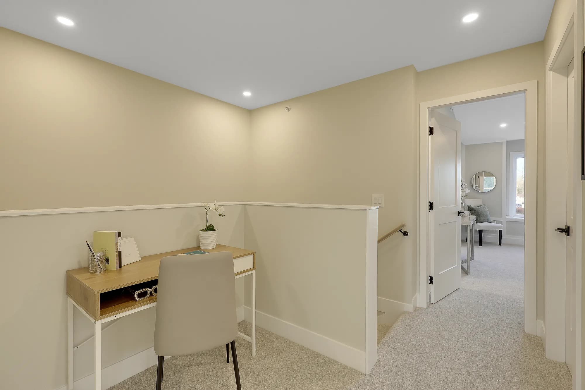 Latimer Landing – An upper-floor hallway landing features a compact wooden work desk with a chair, positioned against neutral beige walls next to a carpeted staircase. Through an open doorway, a glimpse of an adjoining room reveals light-colored carpeting, an accent chair, and a round wall mirror.