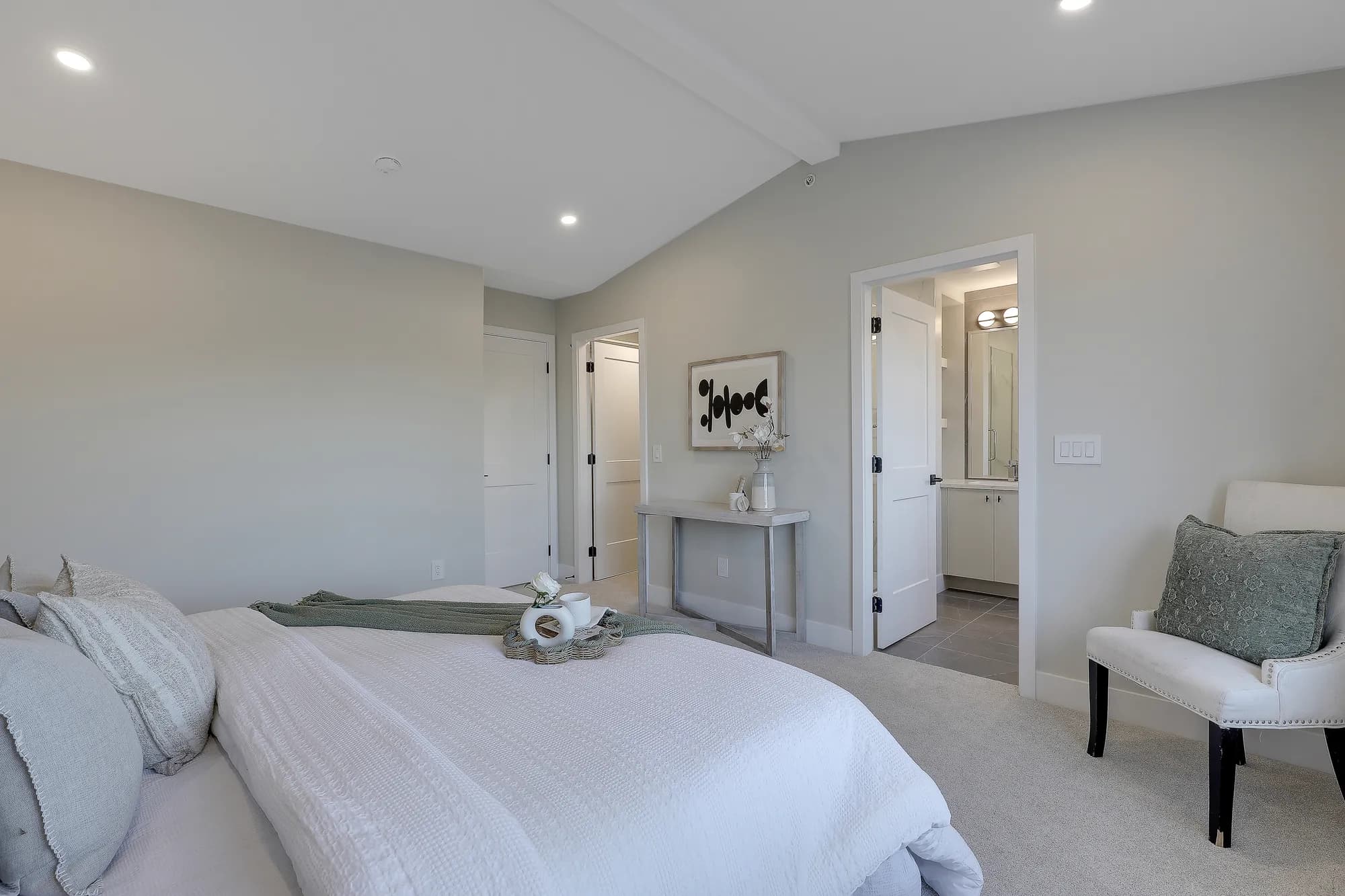 Latimer Landing – A bright, neutral-toned master bedroom featuring a neatly made bed with textured white linens, plush grey and green pillows, and an upholstered accent armchair. An open doorway reveals an en-suite bathroom with tiled floors and a lighted vanity, all under vaulted ceilings with recessed pot lights.