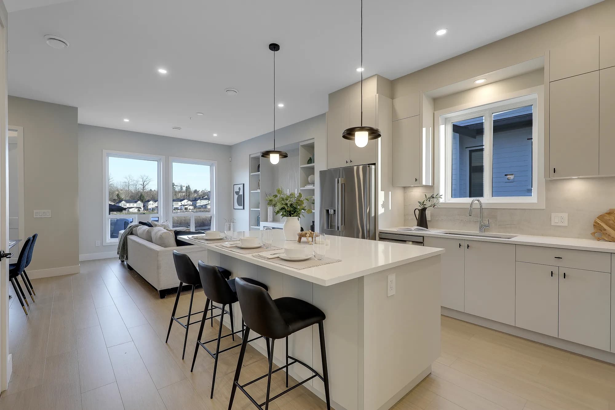 Latimer Landing – A bright, open-concept kitchen and living area featuring a large white quartz island with modern black barstools and pendant lighting overhead. Light wood flooring flows through the space, which includes flat-panel beige cabinetry, stainless steel appliances, and large windows offering views of surrounding homes and trees in daylight.