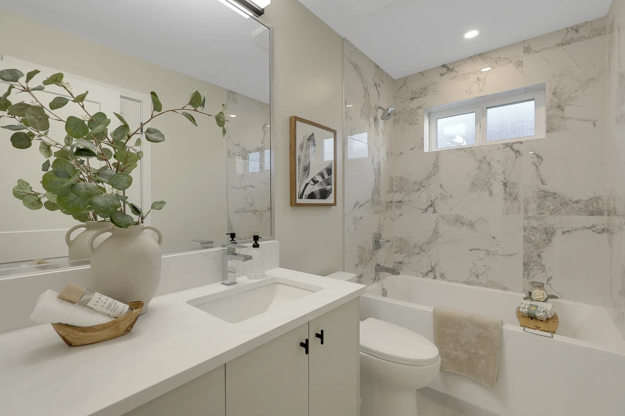 Latimer Landing – A bright, modern bathroom featuring a white vanity with chrome fixtures and a large mirror reflecting marble-patterned wall tiles. In the background, a white bathtub and toilet sit beside tiled walls beneath a small frosted window.