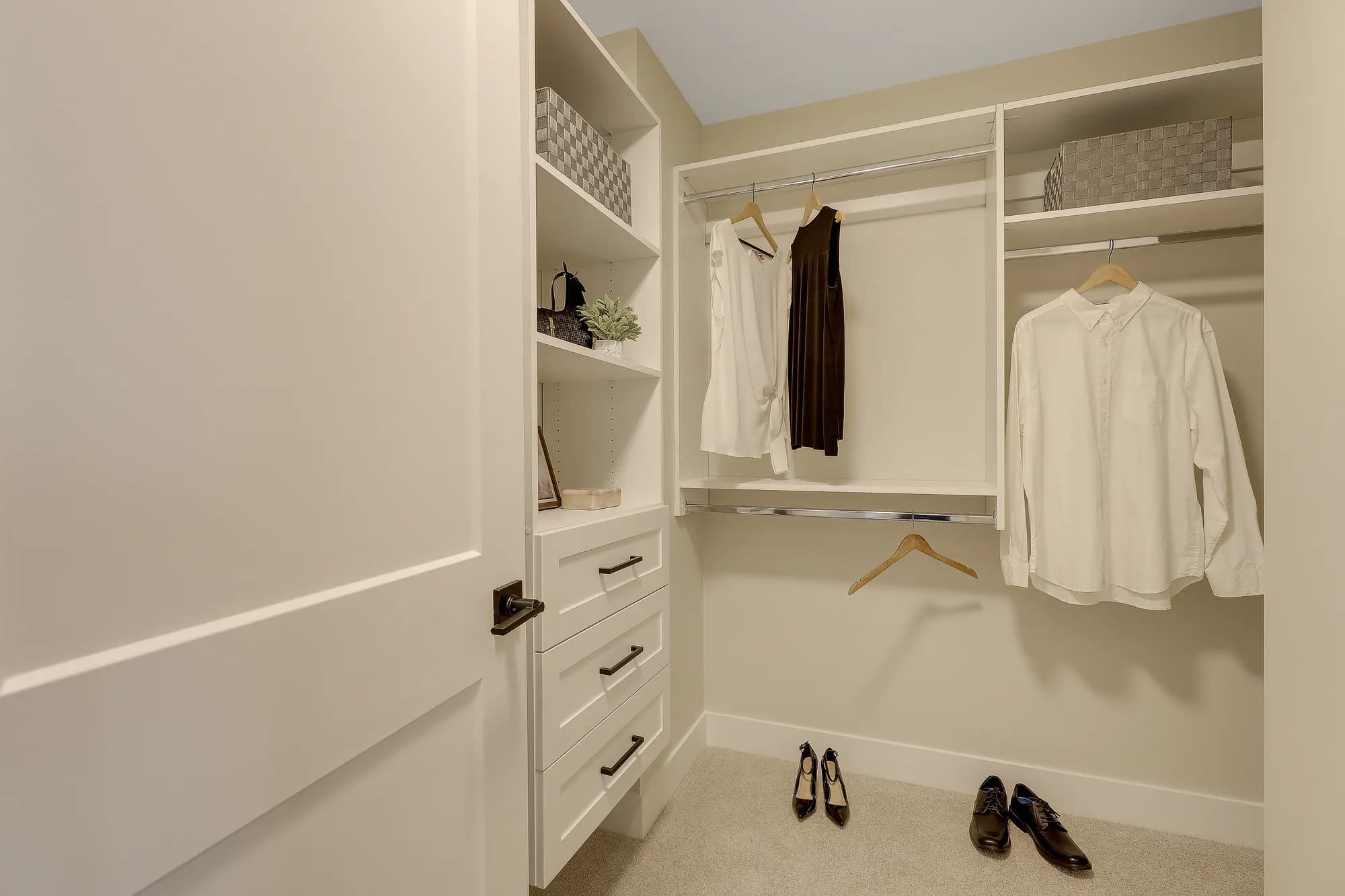 Latimer Landing – A clean, modern walk-in closet featuring white built-in shelving, drawers with black hardware, and multiple chrome hanging rods. The space is lightly staged with a few articles of clothing on wooden hangers, woven storage bins, and dress shoes arranged neatly on the carpeted floor.
