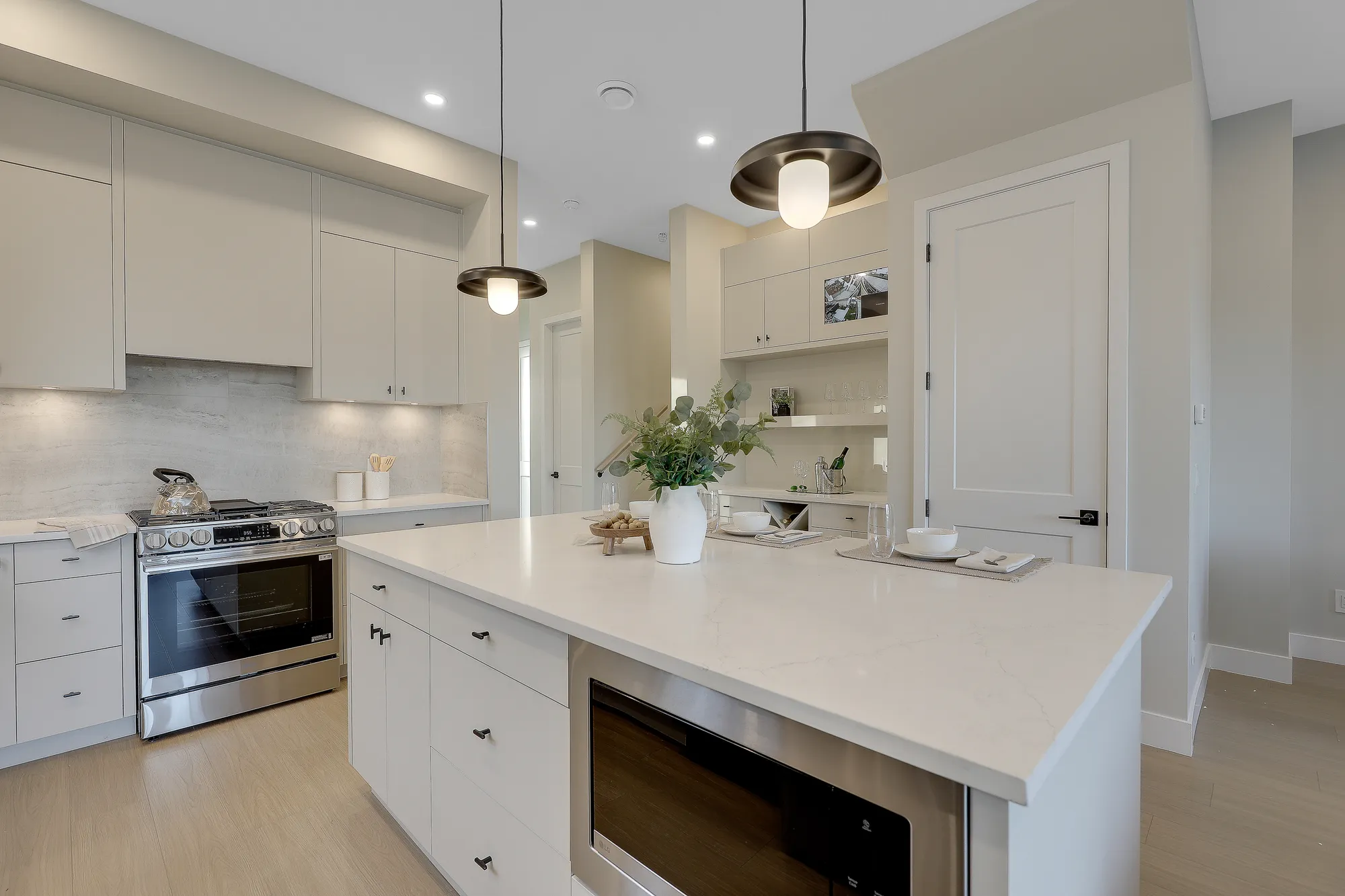 Latimer Landing – A modern, bright kitchen featuring a large white quartz island with an integrated microwave, topped with contemporary pendant lights and dining settings. The space includes sleek off-white flat-panel cabinetry, a stainless steel gas range, light hardwood flooring, and a built-in dry bar or pantry nook in the background.