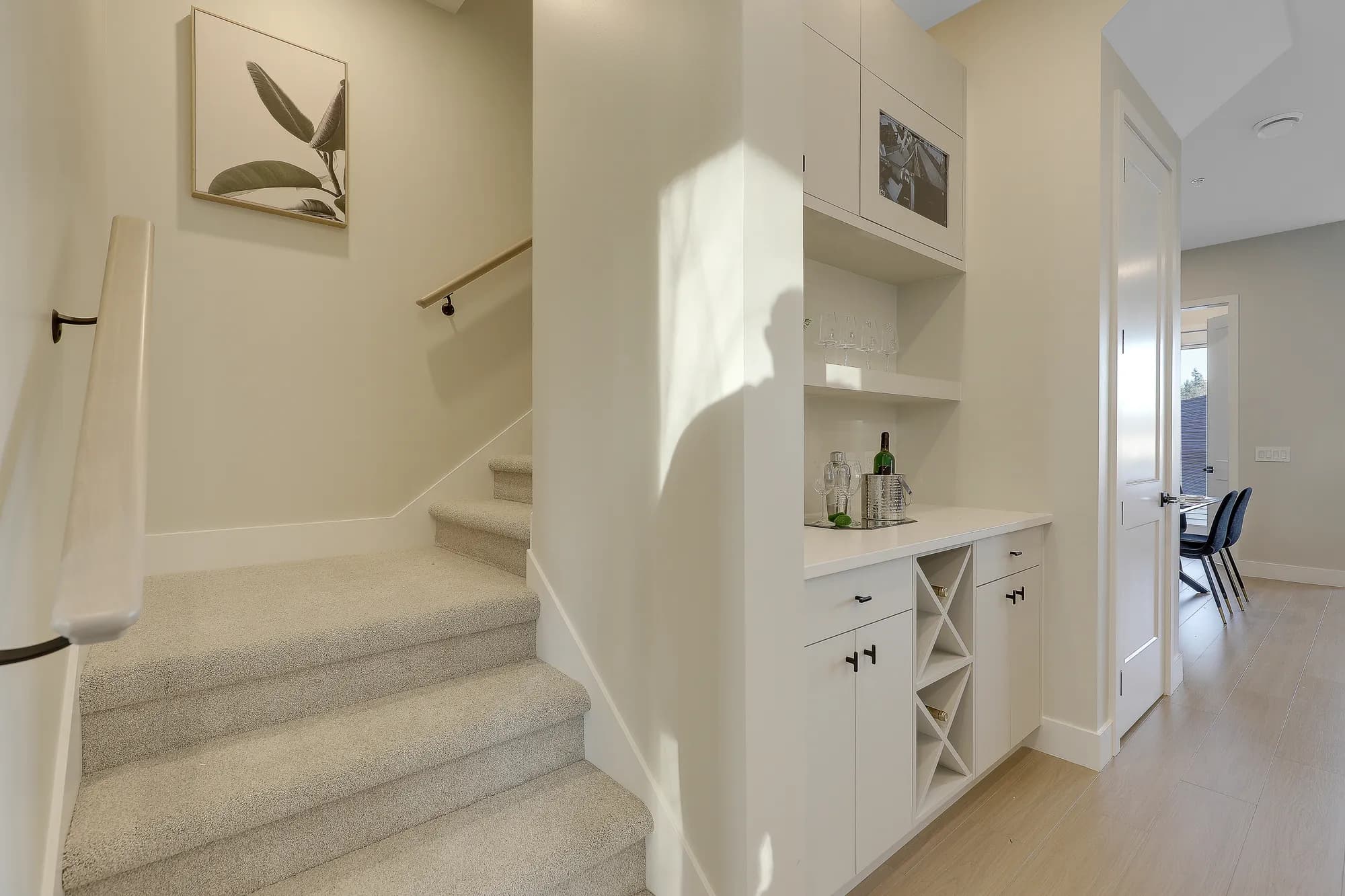 Latimer Landing – A bright residential interior corner showing a carpeted staircase on the left and a custom built-in white dry bar on the right, complete with wine storage and glassware shelves. Sunlight streams across the neutral beige walls and light hardwood flooring, leading into an adjacent dining area in the background.