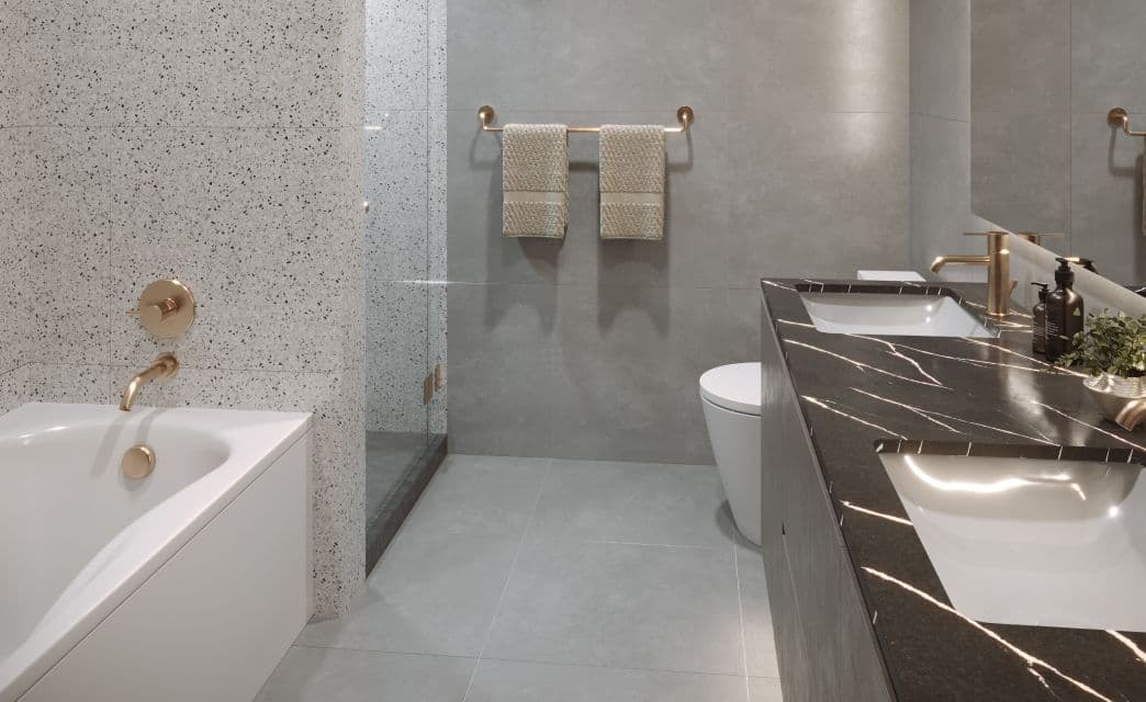 Gryphon Nova Condos – A bathroom rendering with terrazzo-look tile, a soaker tub with gold fixtures, and a dark marble double vanity with gold hardware.