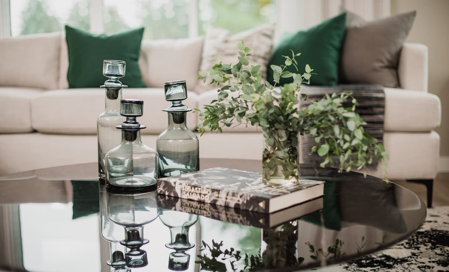 Beaumont – A reflective black glass coffee table is styled with tint-blue glass decanters, a coffee table book, and a vase of fresh eucalyptus branches. In the softly blurred background, a cream-colored sectional sofa features deep green and grey accent pillows, creating an elegant and modern living room ambiance.