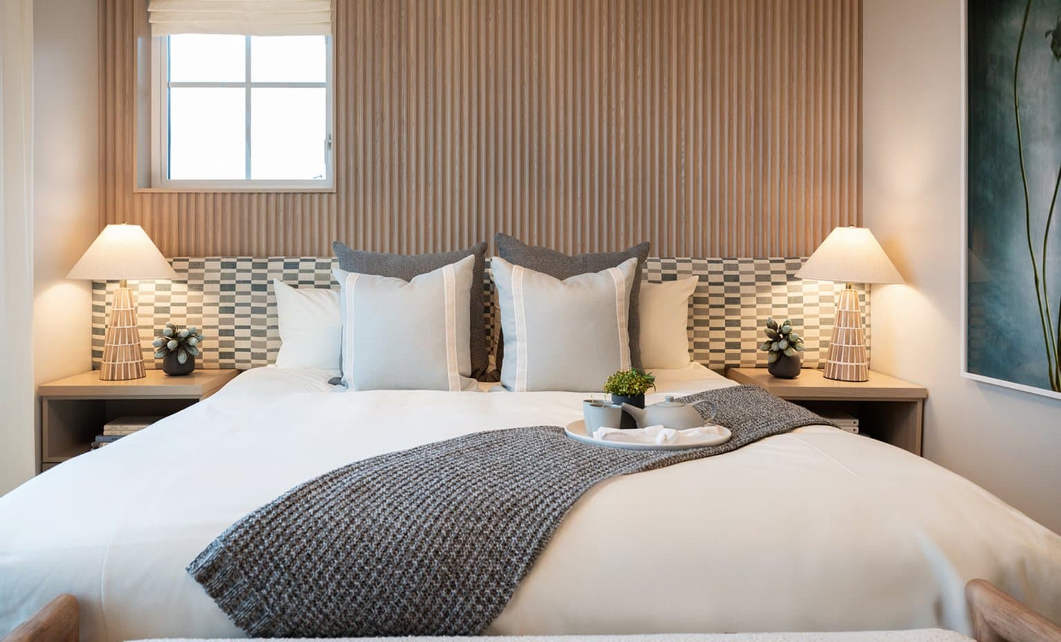 Beaumont – A warmly lit bedroom featuring a bed dressed in crisp white linens, grey throw pillows, and a textured knit runner across the foot. The headboard wall combines vertical wood fluting with a patterned tiled accent, flanked by matching light-wood nightstands with glowing table lamps.