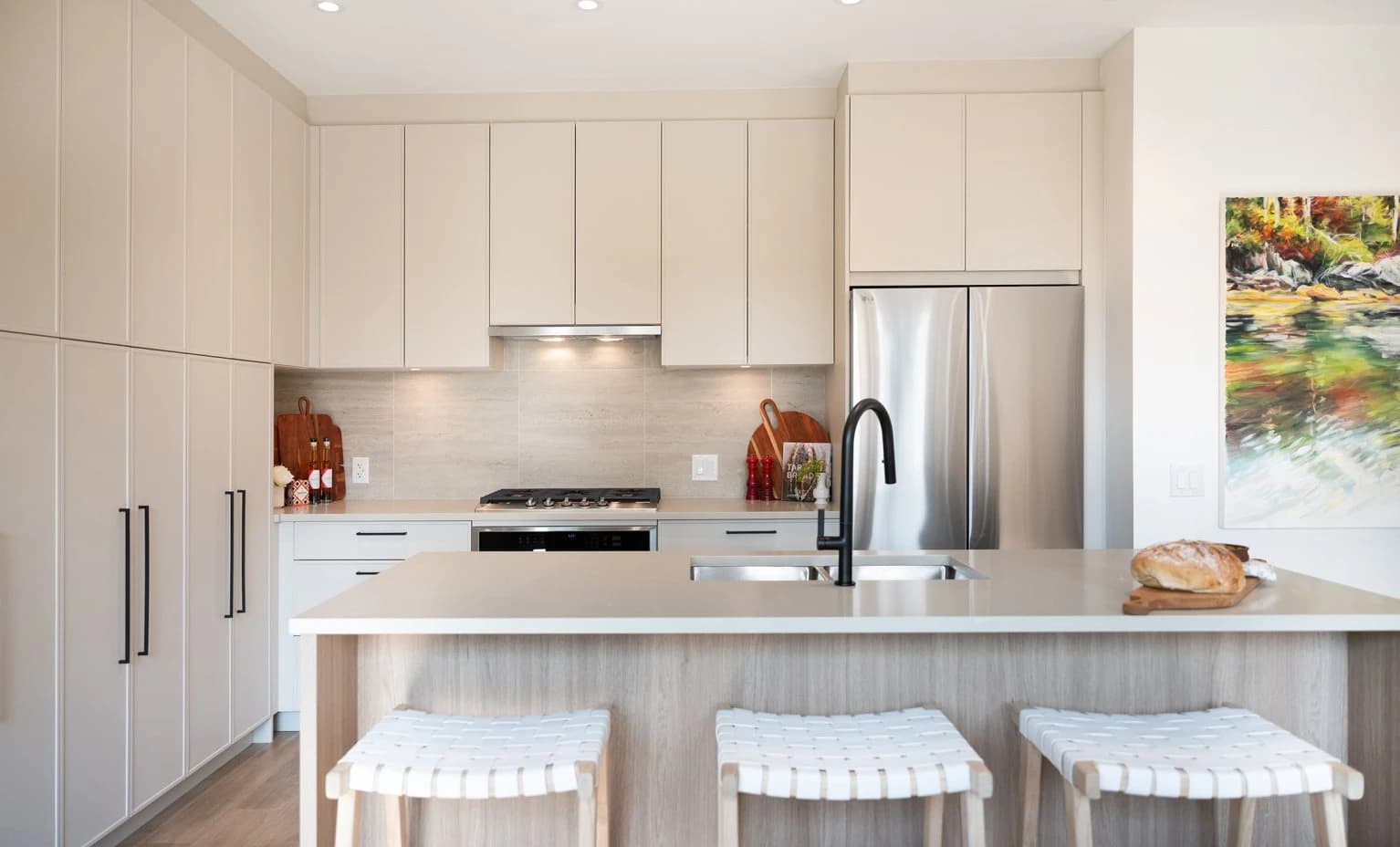 Beaumont – A modern, neutral-toned kitchen featuring light beige flat-panel cabinetry, a light wood kitchen island with woven barstools, and stainless steel appliances. The space is illuminated with recessed lighting and under-cabinet lights, complemented by a matte black gooseneck faucet and a colorful nature painting on the adjacent wall.