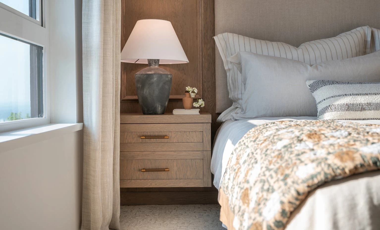Beaumont – A cozy bedroom detail showing a layered, neatly styled bed next to a floating light-wood nightstand with an illuminated table lamp. Soft natural daylight pours in from an adjacent window, highlighting neutral-toned linens, textured pillows, and wood paneling on the wall.
