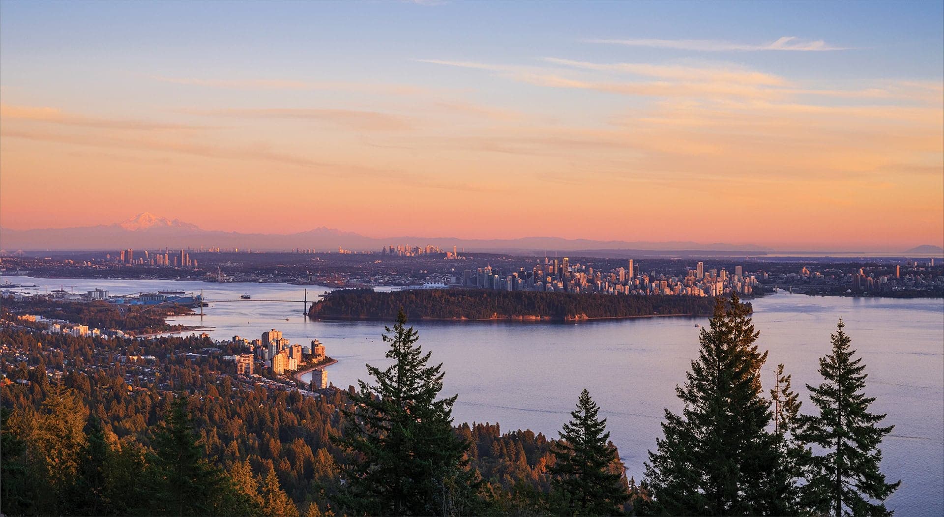 The Summit – A high-elevation panoramic vista looks across evergreen forested hills toward a vast body of water, an urban skyline, and a large forested park peninsula. A suspension bridge spans the inlet and distant snow-capped mountains rise beneath a soft pink and gold twilight sky.
