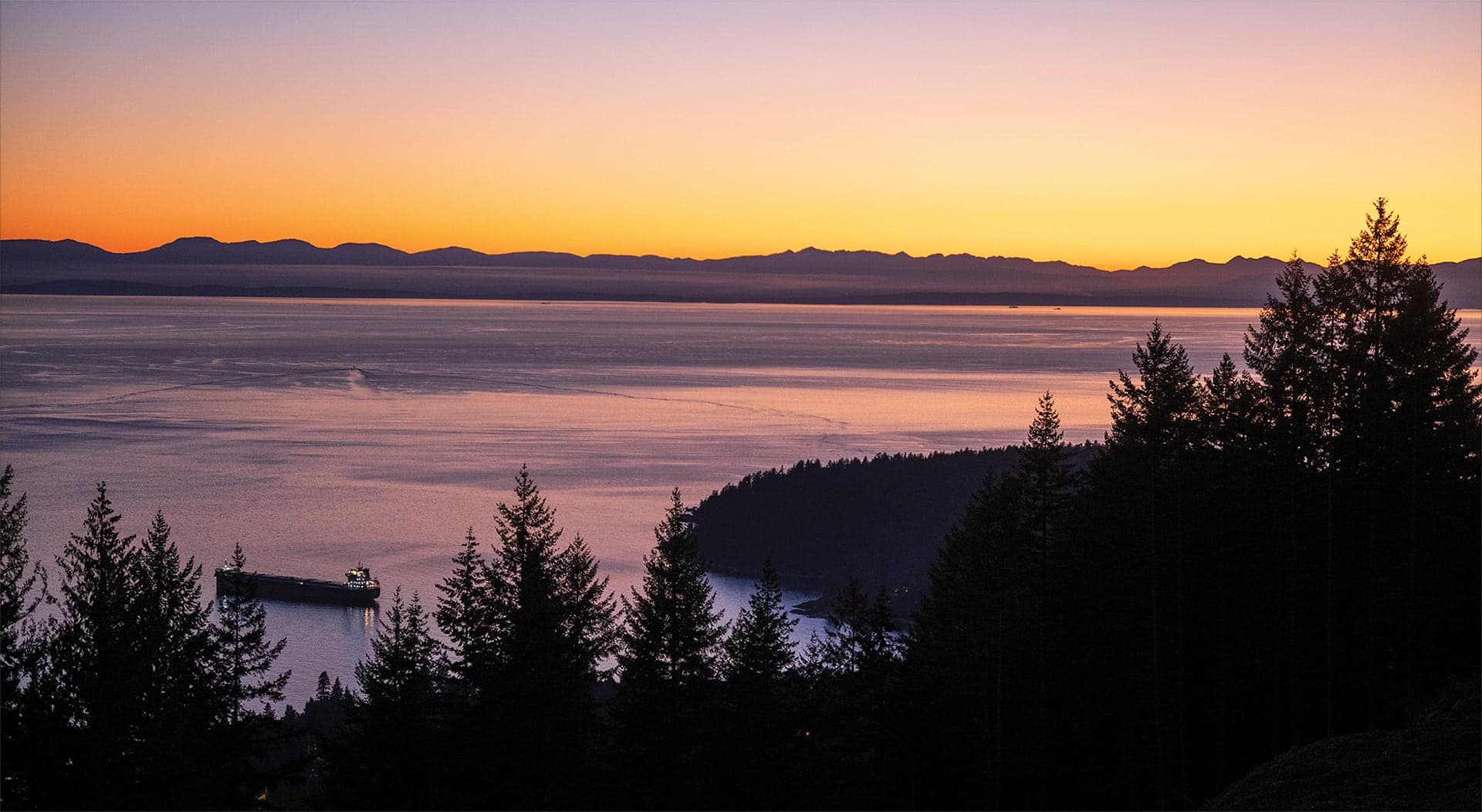 The Summit – A wide scenic sunset view overlooking an expansive ocean bay, framed by dark evergreen treetops in the foreground and a distant mountain range along the horizon. An illuminated cargo ship rests on the water below, under a sky glowing with gradients of vibrant orange, purple, and soft pink.