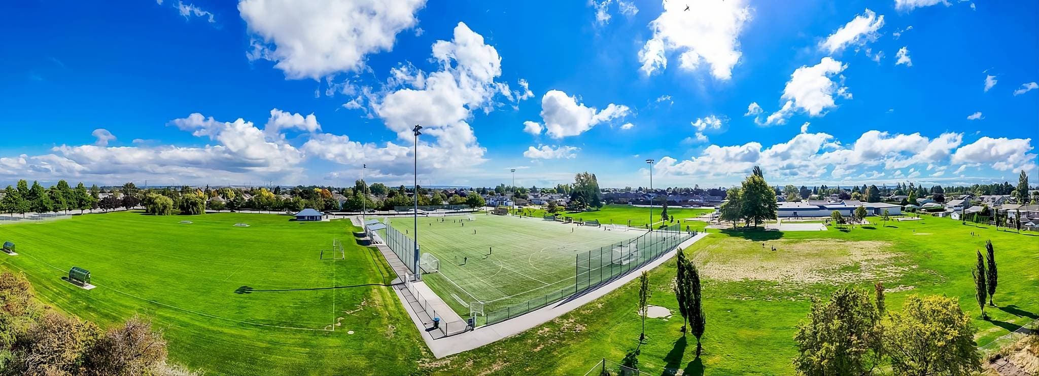 Skye at Holly Park – Panoramic aerial photo of adjacent sports fields and a park with people playing, houses in the distance.
