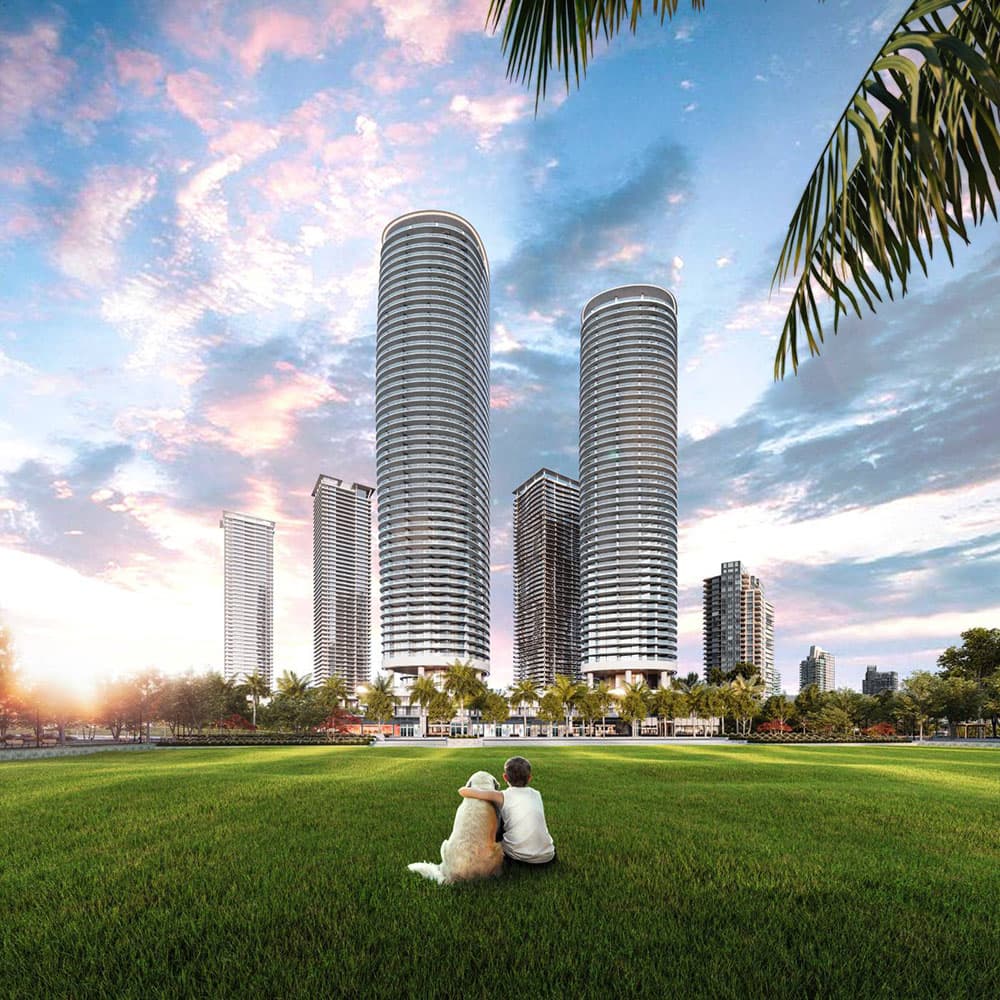 Oasis at Concord Brentwood – A low-angle view shows a child sitting beside a dog on a large green lawn, looking toward two tall, cylindrical glass high-rises lined with wrap-around balconies. Palm trees and additional modern towers frame the background under a vibrant sunset sky filled with pink and blue clouds.