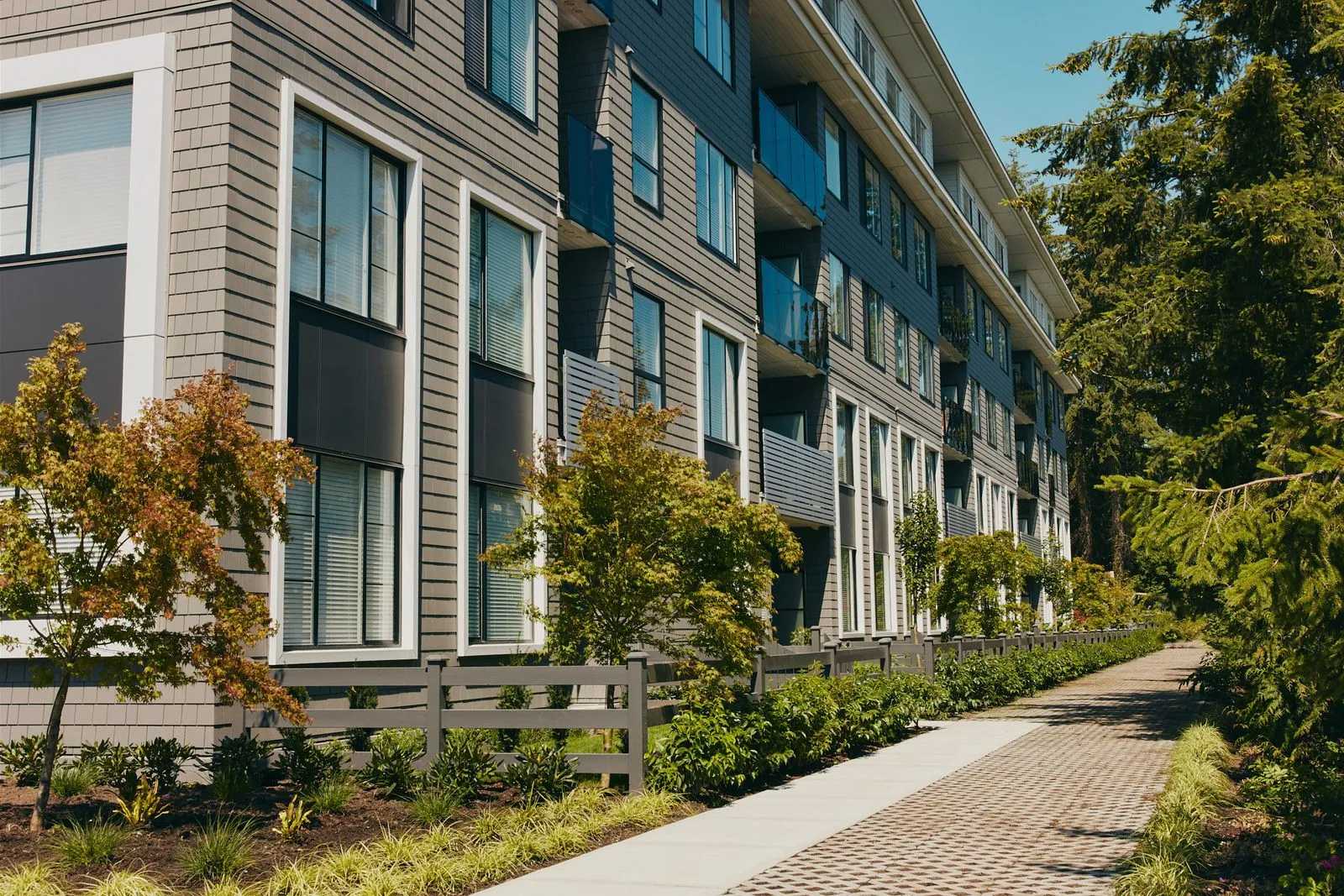 Queens Townhomes – A ground-level view of a modern multi-story residential building featuring grey horizontal siding, dark accent panels, and glass-railed balconies. A paved walkway runs alongside lush landscaping with low fencing, shrubs, and trees under bright daytime sunlight.