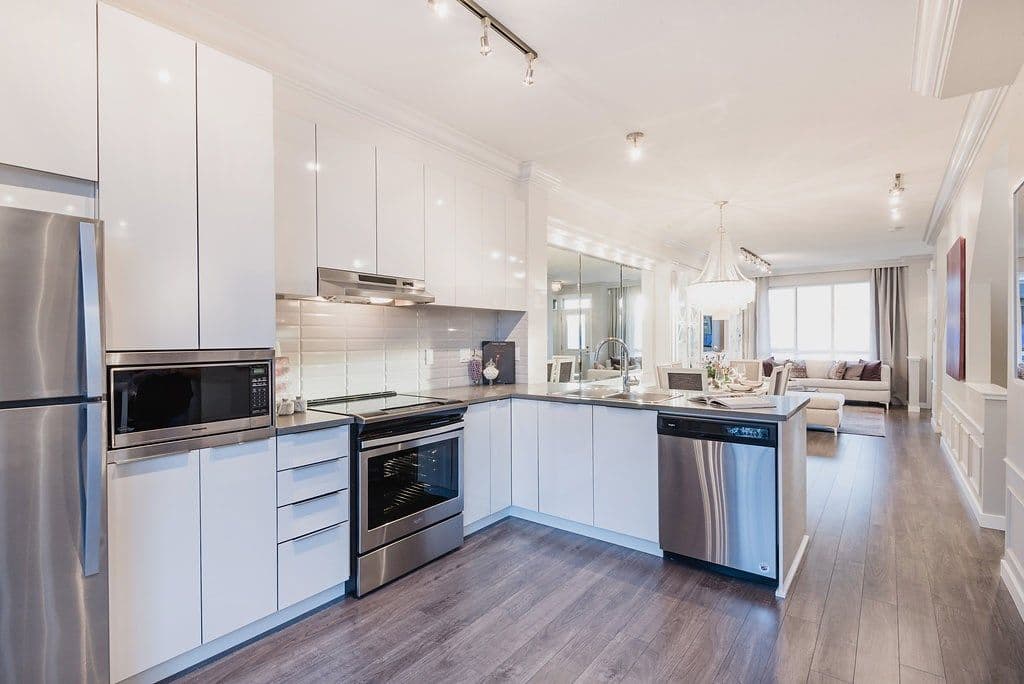 Bristol Heights at Westerleigh – White kitchen with stainless steel appliances, quartz counters, a chandelier over an open dining/living area visible through a mirrored wall.