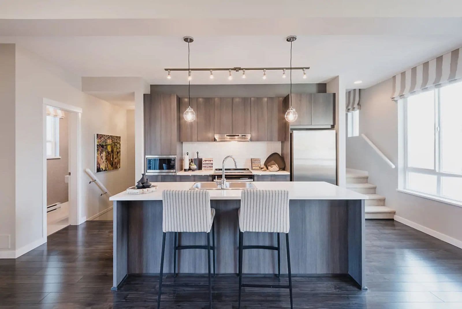 Bristol Heights at Westerleigh – Open-concept kitchen with grey wood-grain cabinets, a large island with bar stools, pendant lighting, and a staircase visible in the background.