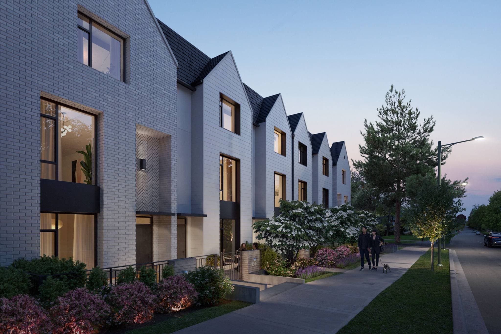 Whitford – A row of modern three-story townhomes with white brick and light gray siding features prominent gabled roofs and large black-framed windows at dusk. A paved sidewalk lined with flowering bushes, small trees, and illuminated streetlights runs past the entryways where residents walk along a quiet street.