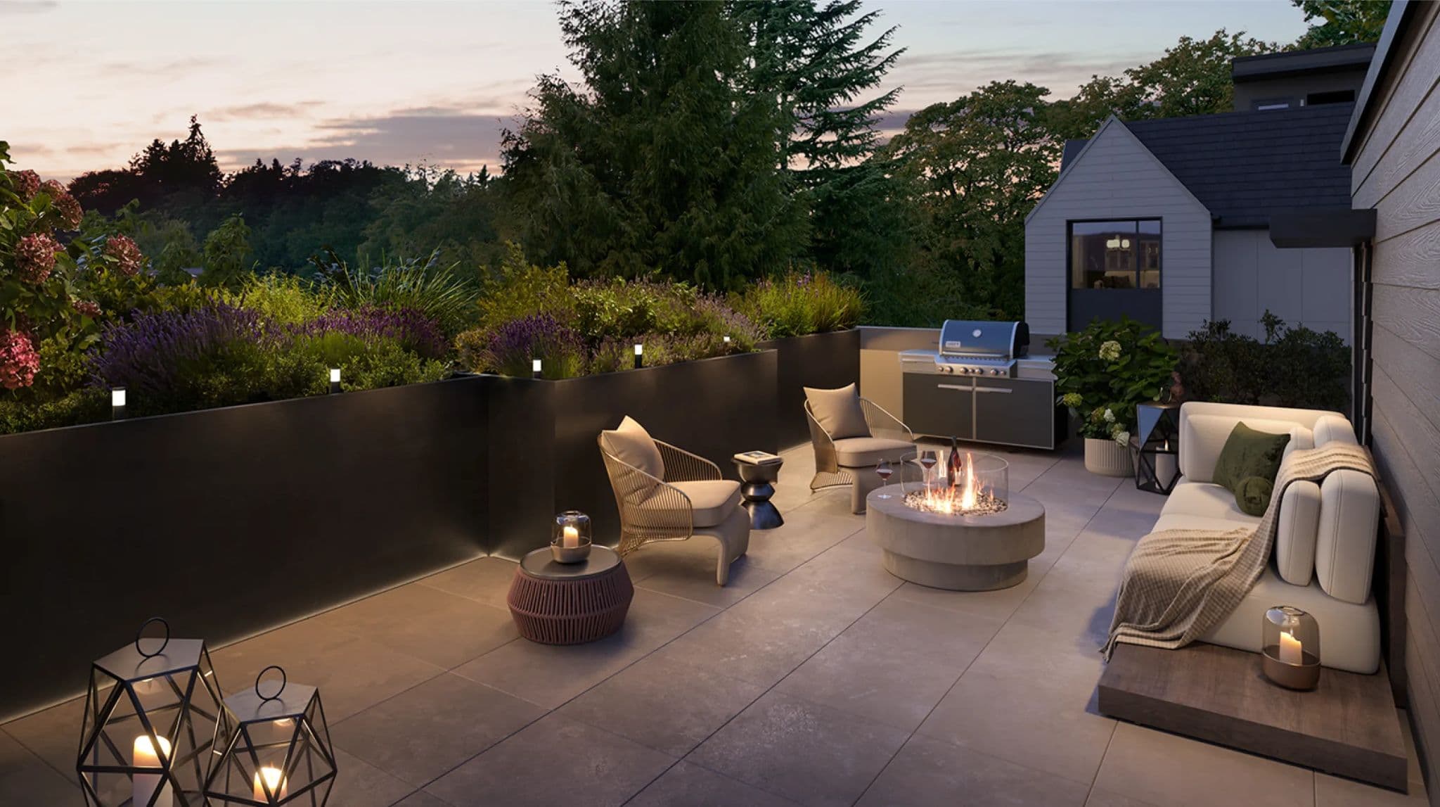 Whitford – A twilight view of a cozy rooftop patio furnished with a plush outdoor sofa, armchairs, a glowing concrete fire pit, and an outdoor BBQ station. Deep planter boxes filled with lush greenery and lavender border the tiled terrace, overlooking a dense tree line under a soft sunset sky.