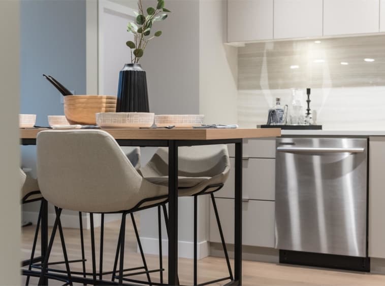 Crescent Court – Dining nook with a wood table set for a meal, beige upholstered chairs, and a stainless steel dishwasher visible in the background kitchen.