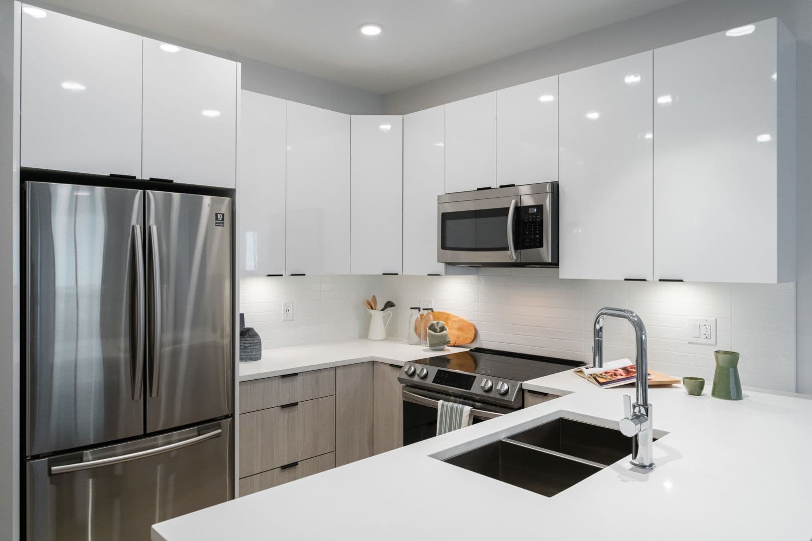 Galilea – A modern, bright kitchen featuring high-gloss white upper cabinets, light wood grain lower drawers, and white quartz countertops with an undermount double sink and chrome faucet. Stainless steel appliances, including a french door refrigerator, over-the-range microwave, and glass-top stove, complete the clean and contemporary layout.