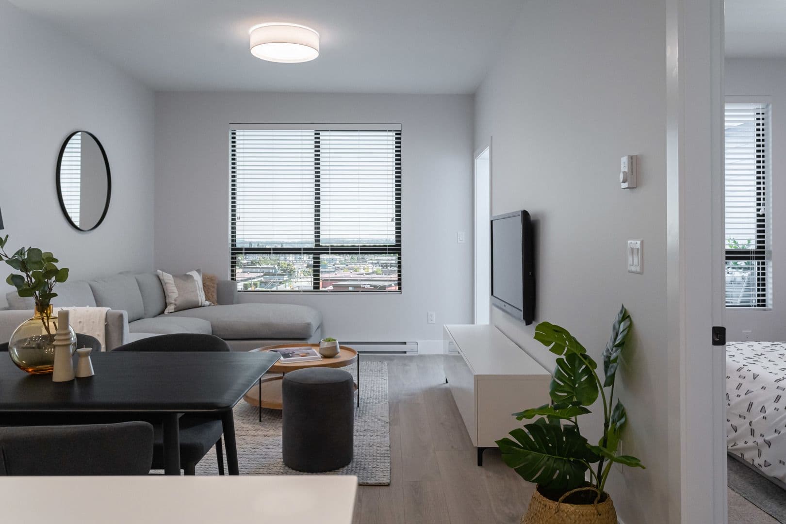 Galilea – A modern open-concept living room featuring a light grey sectional couch, dark dining table, wooden coffee table, and flat-screen TV mounted above a sleek white console. Light wood-look flooring and bright white walls create a clean, airy atmosphere, complimented by natural light streaming through blinds on a large window.