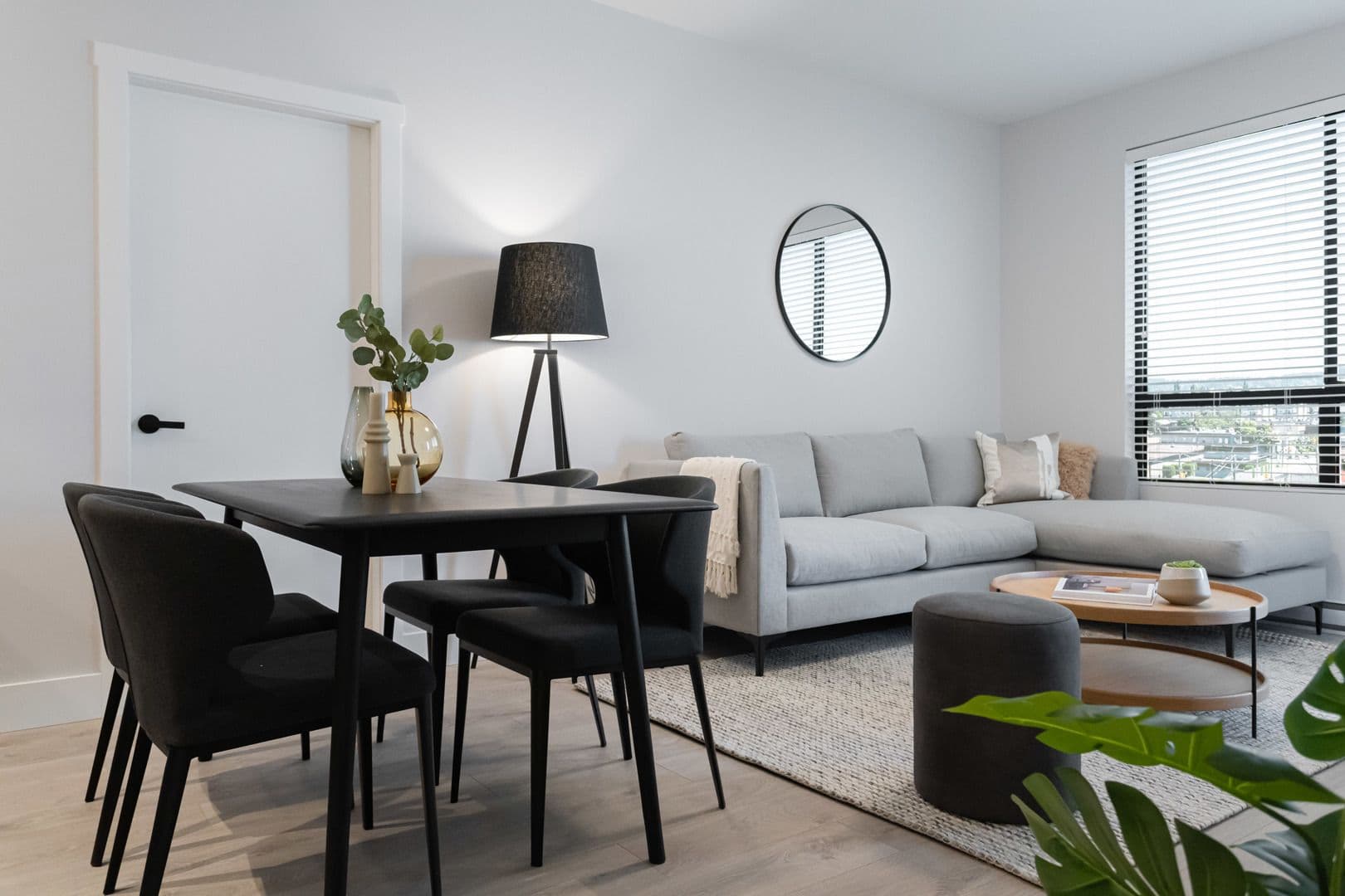 Galilea – A modern open-concept living and dining area featuring light wood flooring, bright white walls, a dark wood dining table set, and a light gray sectional sofa. The brightly lit space is styled with a circular wall mirror, a black tripod floor lamp, a textured area rug, and large windows with horizontal blinds that let in natural daylight.