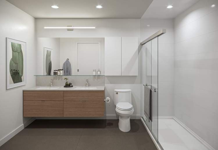One Shaughnessy – Bathroom with a floating wood double vanity, large mirror, toilet, and a glass-enclosed shower with sliding door.