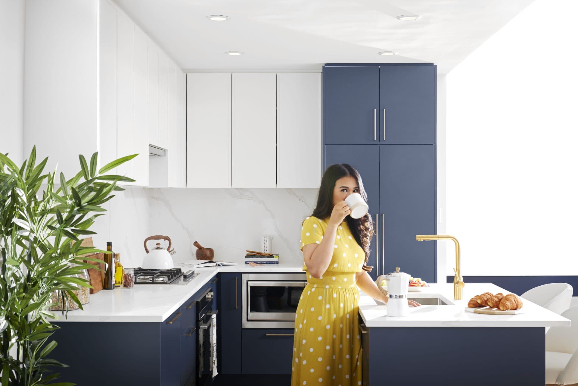 Florin – Lifestyle photo of a woman in a yellow polka-dot dress drinking coffee in a kitchen with navy blue cabinetry, white marble counters, and a brass faucet.