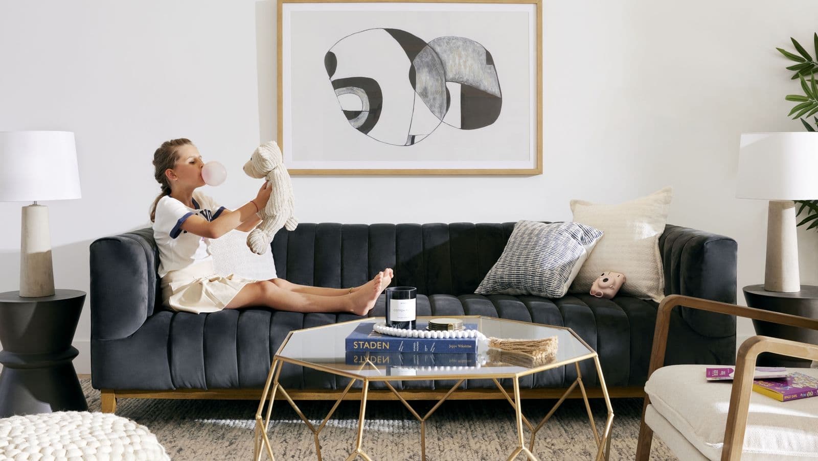 Florin – Lifestyle photo of a girl sitting on a dark blue channel-tufted sofa holding a stuffed toy, with abstract art on the wall and a glass coffee table in front.