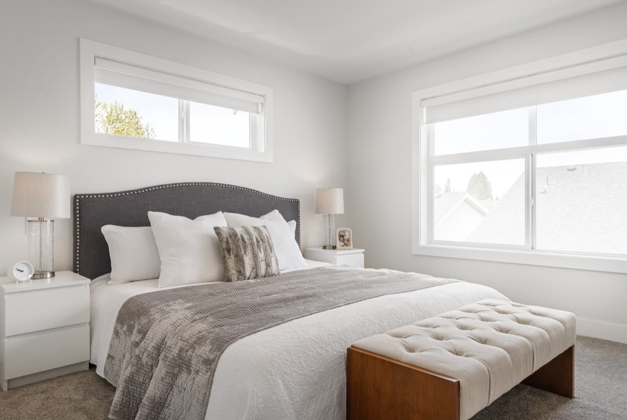 Gordon Square – Bedroom with a grey nailhead-trim headboard, white and grey bedding, and two large windows showing trees and neighboring rooftops.