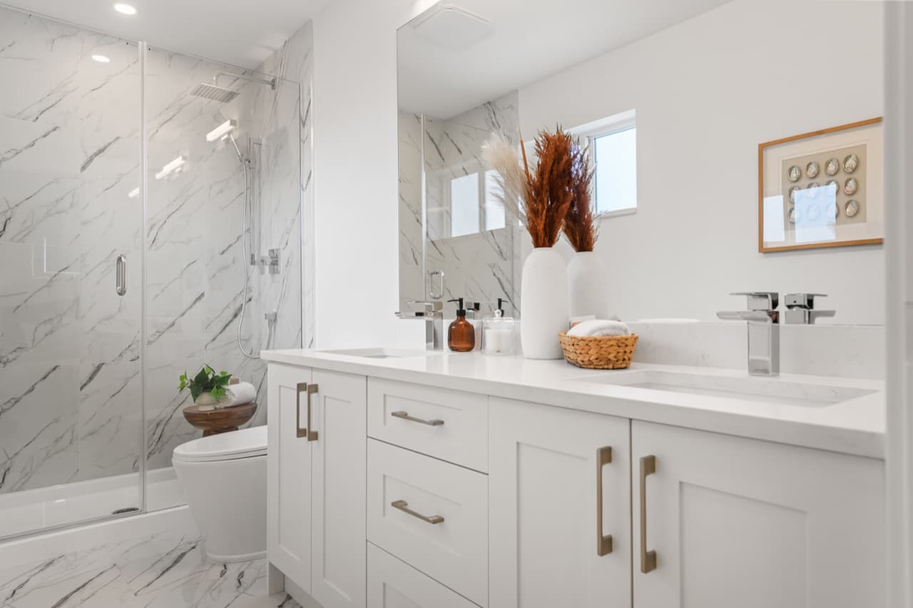 Gordon Square – Bathroom with a marble-tiled glass shower enclosure, toilet, and a white vanity with dried floral arrangements and framed art.