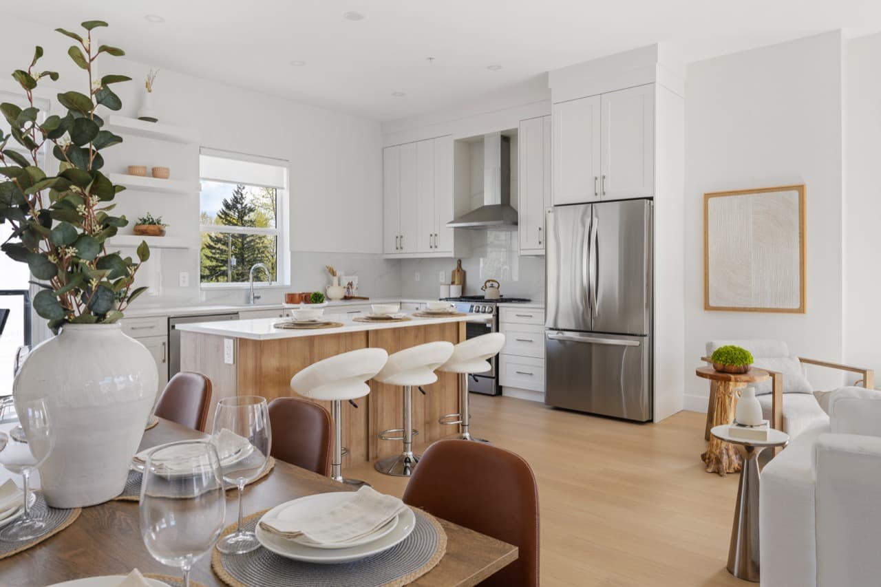 Gordon Square – Open-concept kitchen with white cabinetry, a wood-tone island, stainless fridge, and a dining table with a large potted plant in the foreground.