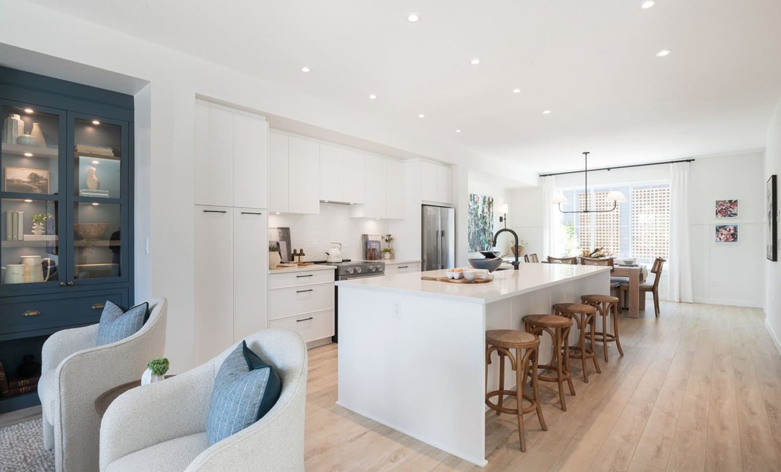 Landon Gate – A bright, open-concept modern kitchen features a large white island with wooden barstools, light flooring, and white cabinetry complemented by a dark blue built-in display cabinet. In the foreground sit two light-colored armchairs, while a sunny dining area with large windows is visible in the background.