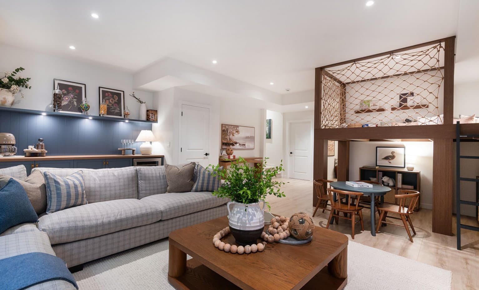Landon Gate – A cozy family basement or play area featuring a large plaid sectional sofa paired with a square wooden coffee table in the foreground. In the background, blue built-in cabinetry sits under a display shelf, while a wooden loft structure with rope netting creates a dedicated play nook above a small child-sized table and chairs set.