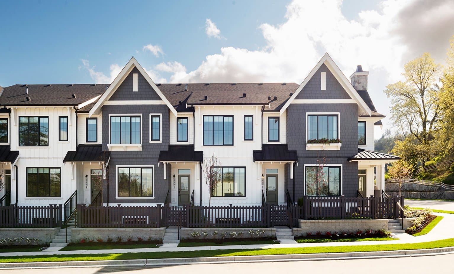 Landon Gate – A row of modern townhomes featuring dark gray shingle siding paired with white board-and-batten accents, black-framed windows, and gabled rooflines. The street front includes small fenced yards with manicured landscaping, concrete steps, and a sidewalk under a bright, partly cloudy sky.