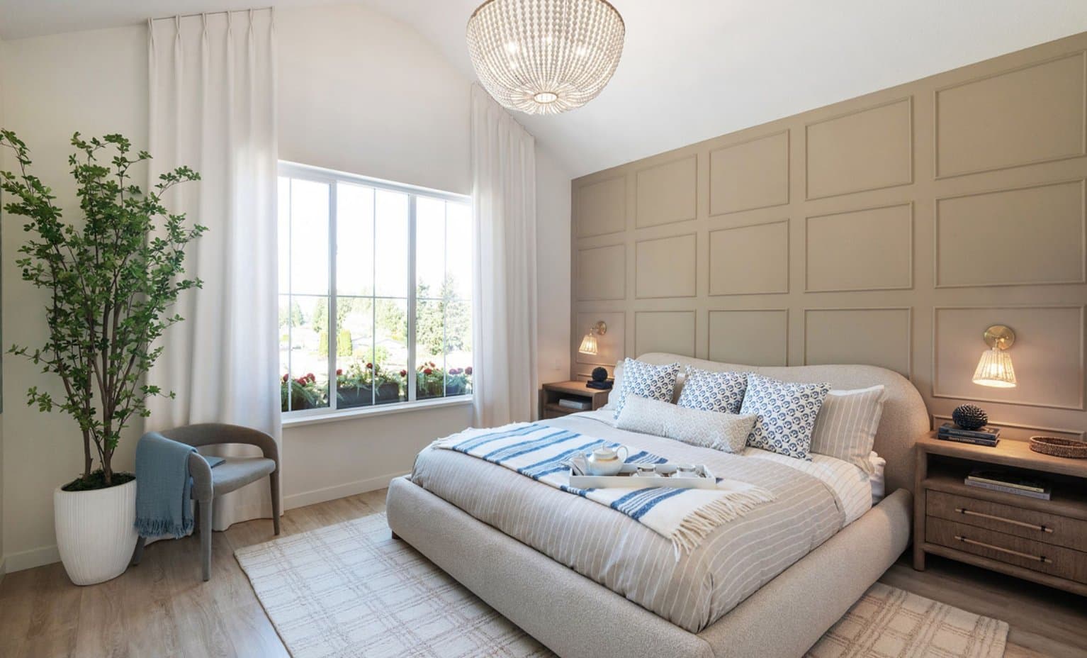 Landon Gate – A bright, elegant bedroom featuring a bed with a light beige upholstered frame, blue accent pillows, and a wooden tray set on top. The room is decorated with a beige grid feature wall, a spherical beaded chandelier, wall sconces over nightstands, and a large window draped in white curtains that overlooks greenery.
