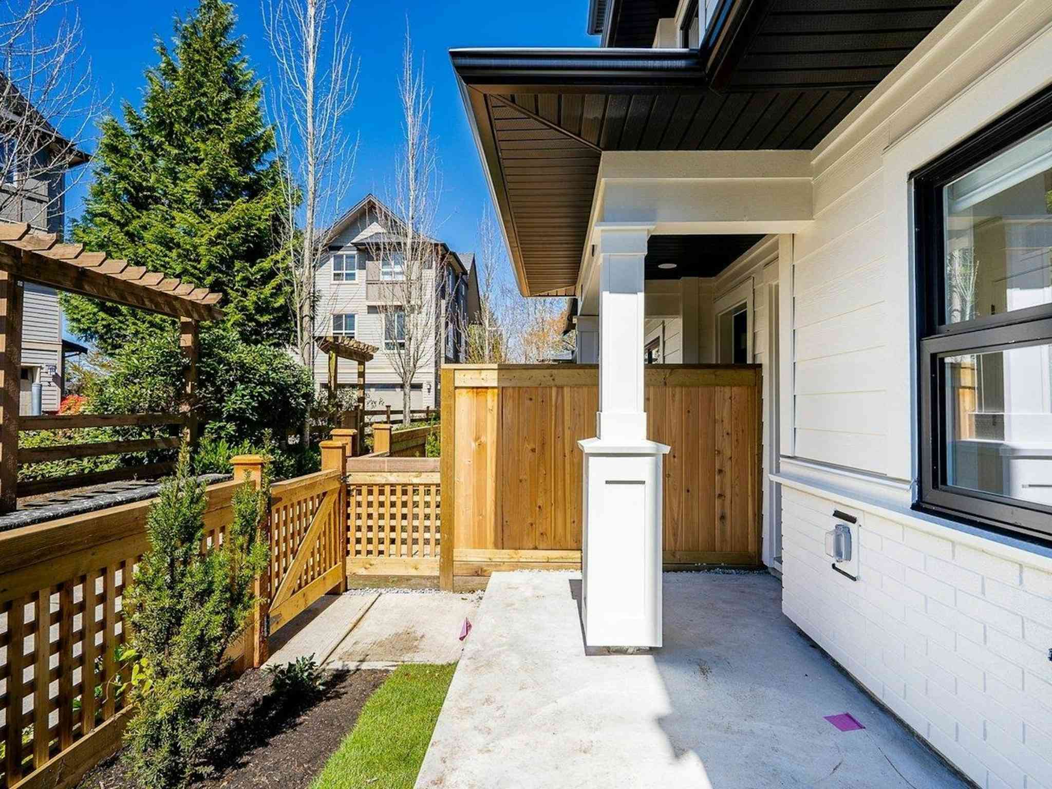 Hyde Park – A ground-level view of a townhouse's covered front porch featuring white siding, a supportive pillar, and a concrete patio. The yard is enclosed by a natural wood fence and lattice gate, with neighboring townhomes and lush evergreen trees visible under a bright, sunny blue sky.