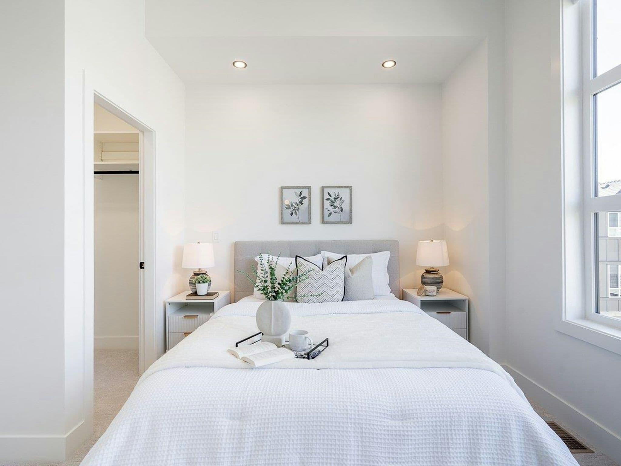 Hyde Park – A bright, contemporary bedroom features a neatly made bed with white waffle-knit bedding, neutral accent pillows, and matching nightstands with soft table lamps. High ceilings with recessed lighting, an open walk-in closet to the left, and a tall window to the right bring in ample natural daylight.