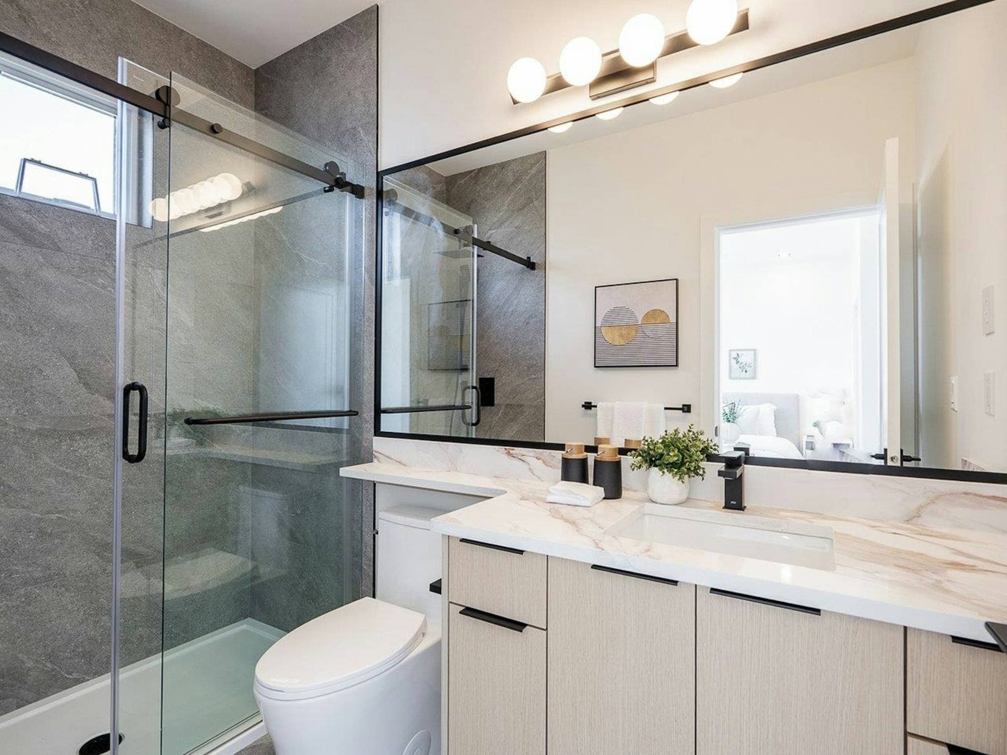 Hyde Park – A modern ensuite bathroom featuring a light wood vanity with a white marble-patterned countertop, matte black hardware, and an expansive framed mirror illuminated by vanity bulb lighting. A glass-enclosed walk-in shower with large-format grey stone tiles sits adjacent to the toilet, reflecting a softly lit contemporary interior.