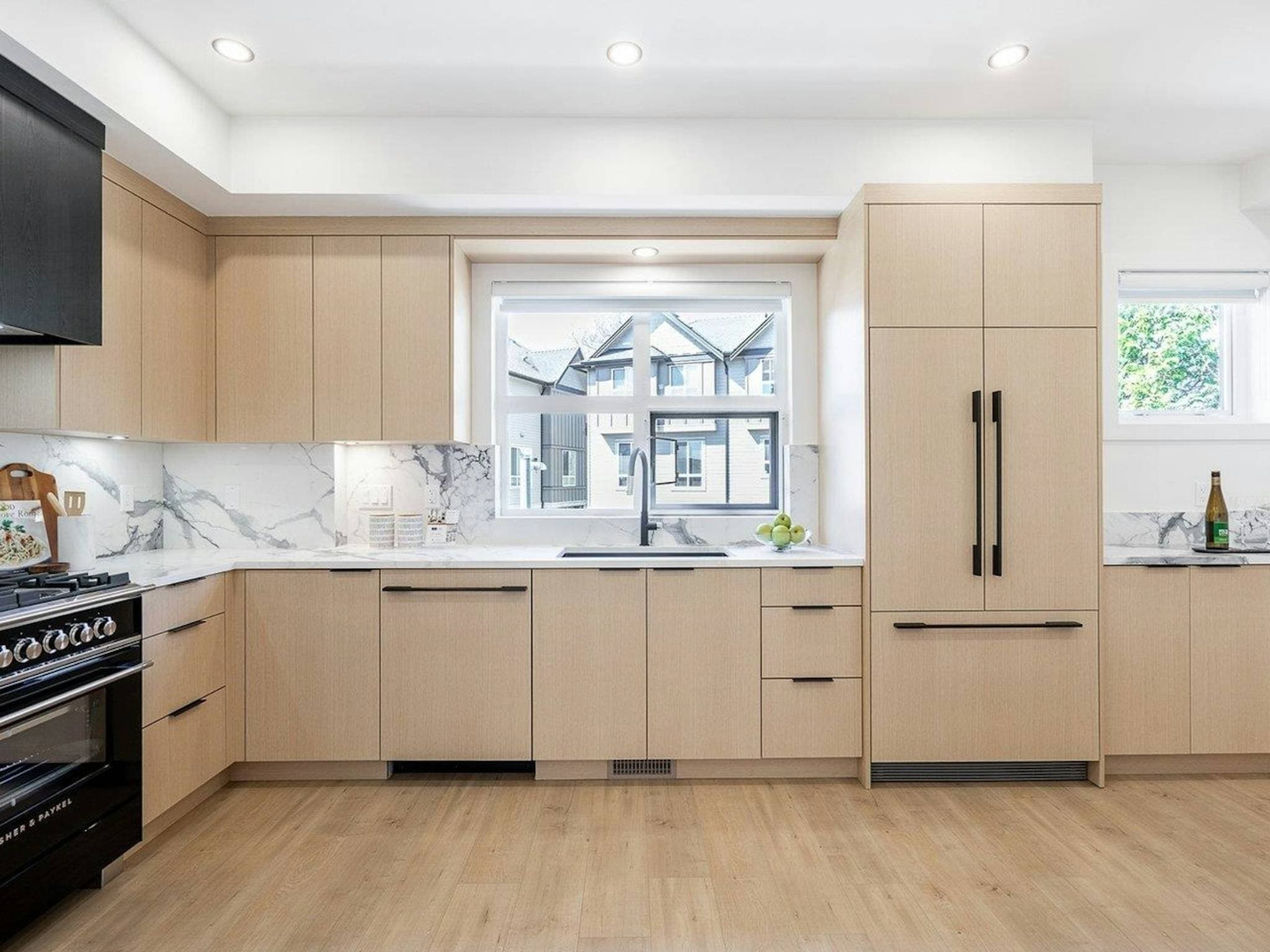 Hyde Park – A modern kitchen featuring flat-panel light wood cabinetry, integrated appliances, black hardware, and white marble-patterned countertops with a matching full-height backsplash. A large picture window above the undermount sink lets in bright natural daylight, offering a view of nearby residential townhomes.