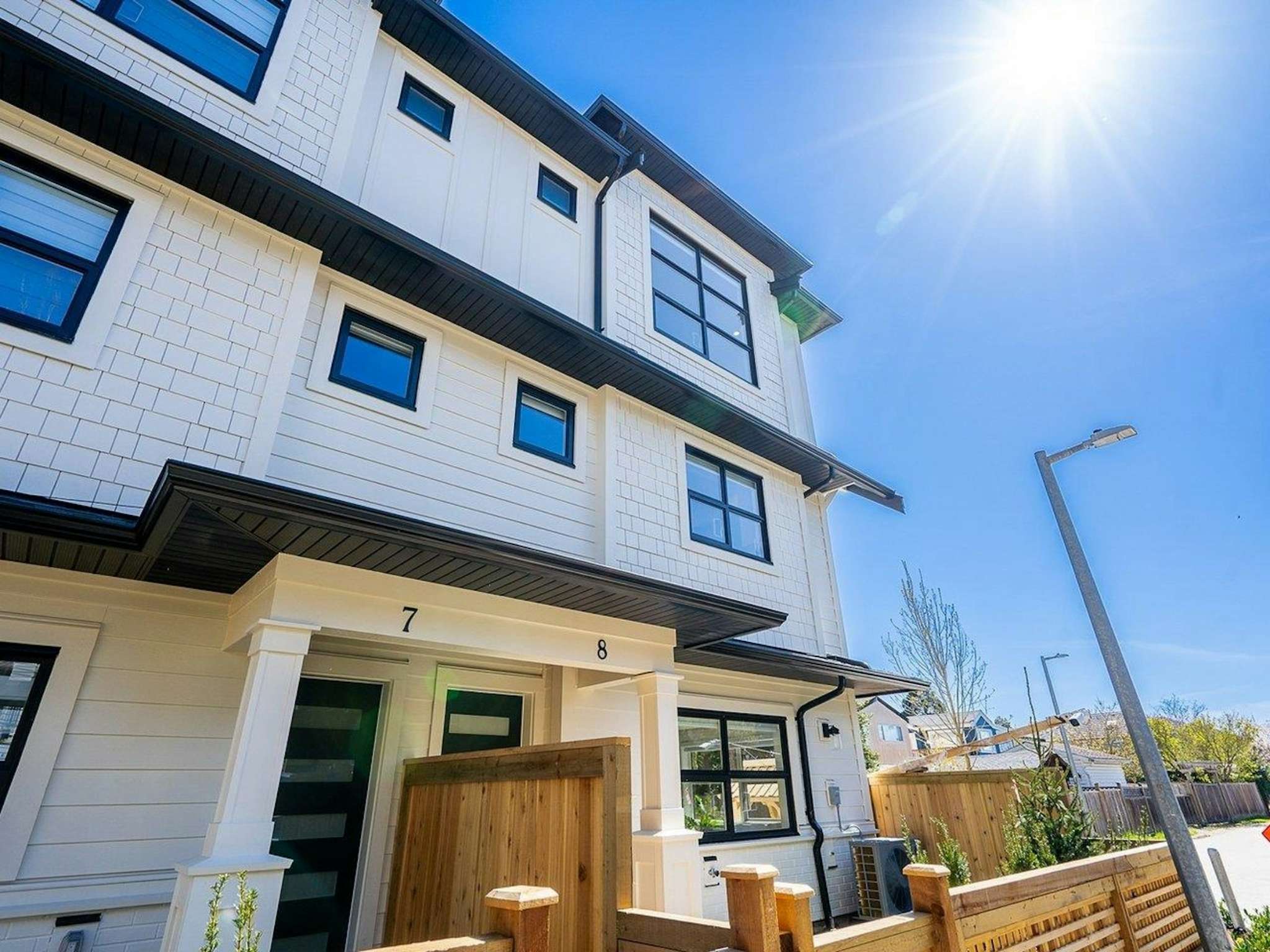 Hyde Park – A low-angle exterior shot of a modern multi-story townhouse featuring white siding and shake paneling, black-trimmed windows, and wood-fenced entry courtyards. The building is framed against a clear bright blue sky with a prominent sun flare above the roofline.
