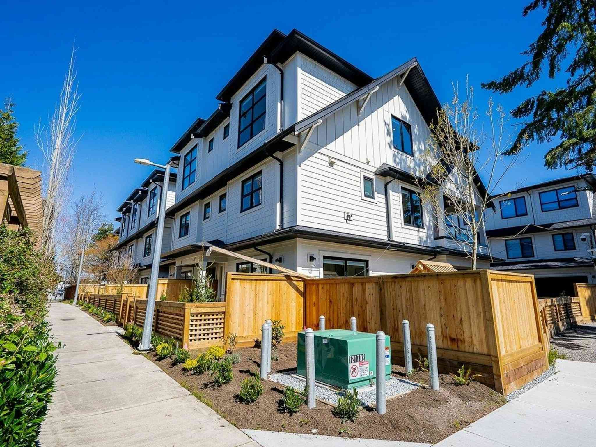 Hyde Park – A ground-level view of modern multi-story townhomes featuring crisp white siding with dark trim and gabled roofs under a clear blue sky. Wooden privacy fencing lines the front yards along a concrete sidewalk, with young landscaping and utility boxes visible in the foreground.