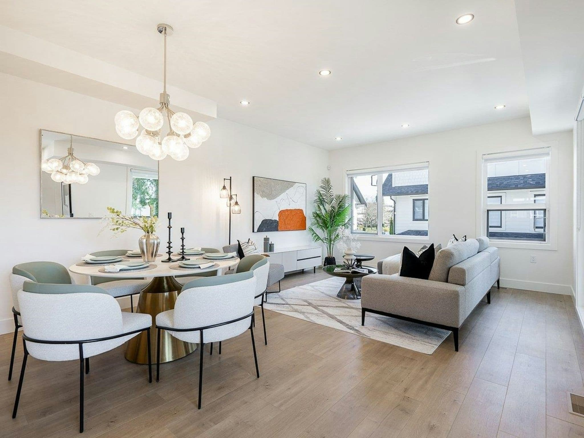 Hyde Park – A bright, open-concept dining and living area featuring light wood flooring, white walls, and contemporary furnishings. A modern globe chandelier hangs above a round dining table with seating for four, adjacent to a living space furnished with a gray sofa, abstract wall art, and windows that look out onto neighboring homes.