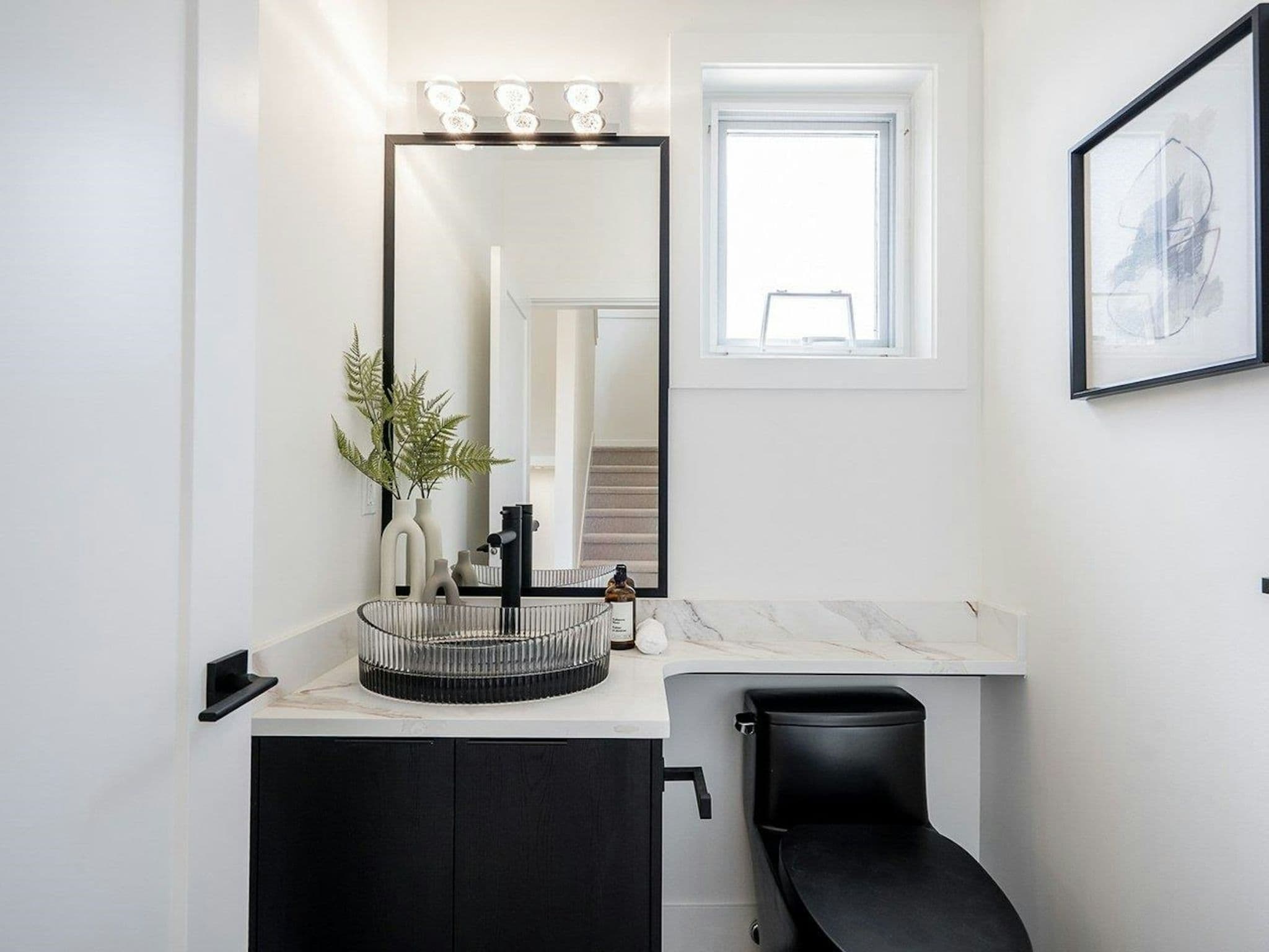Hyde Park – A modern powder room featuring a dark vanity with a ribbed glass vessel sink, matte black hardware, and an extended white marble countertop over a sleek black toilet. A large black-framed vertical mirror and a small frosted window reflect bright daylight into the crisp white space.