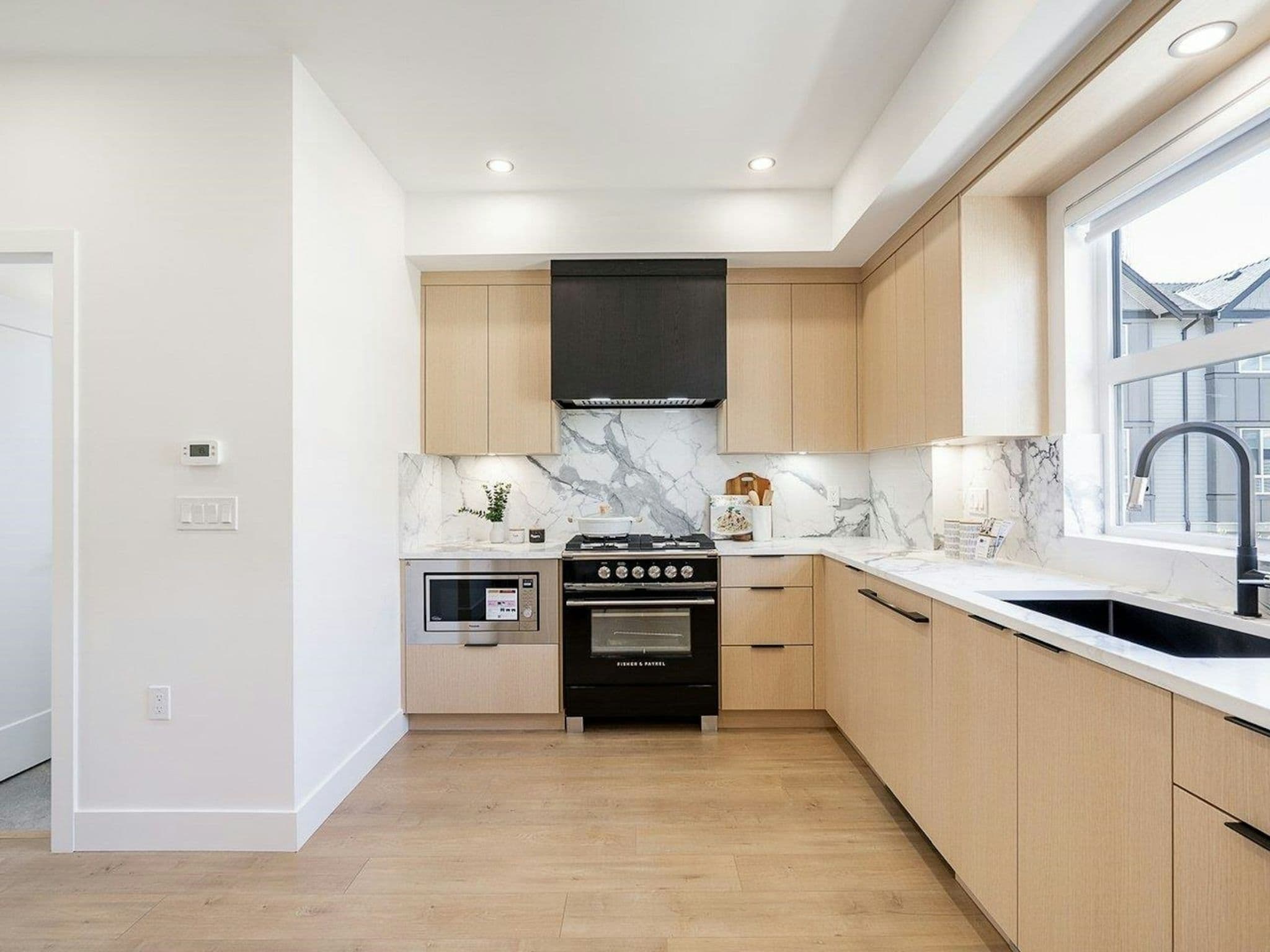Hyde Park – A modern corner kitchen featuring light wood flat-panel cabinetry, white marble-patterned countertops, and a matching full-height backsplash. A bold black range and matching black range hood anchor the center of the space, complemented by light wood flooring and a sink placed beneath a bright side window.