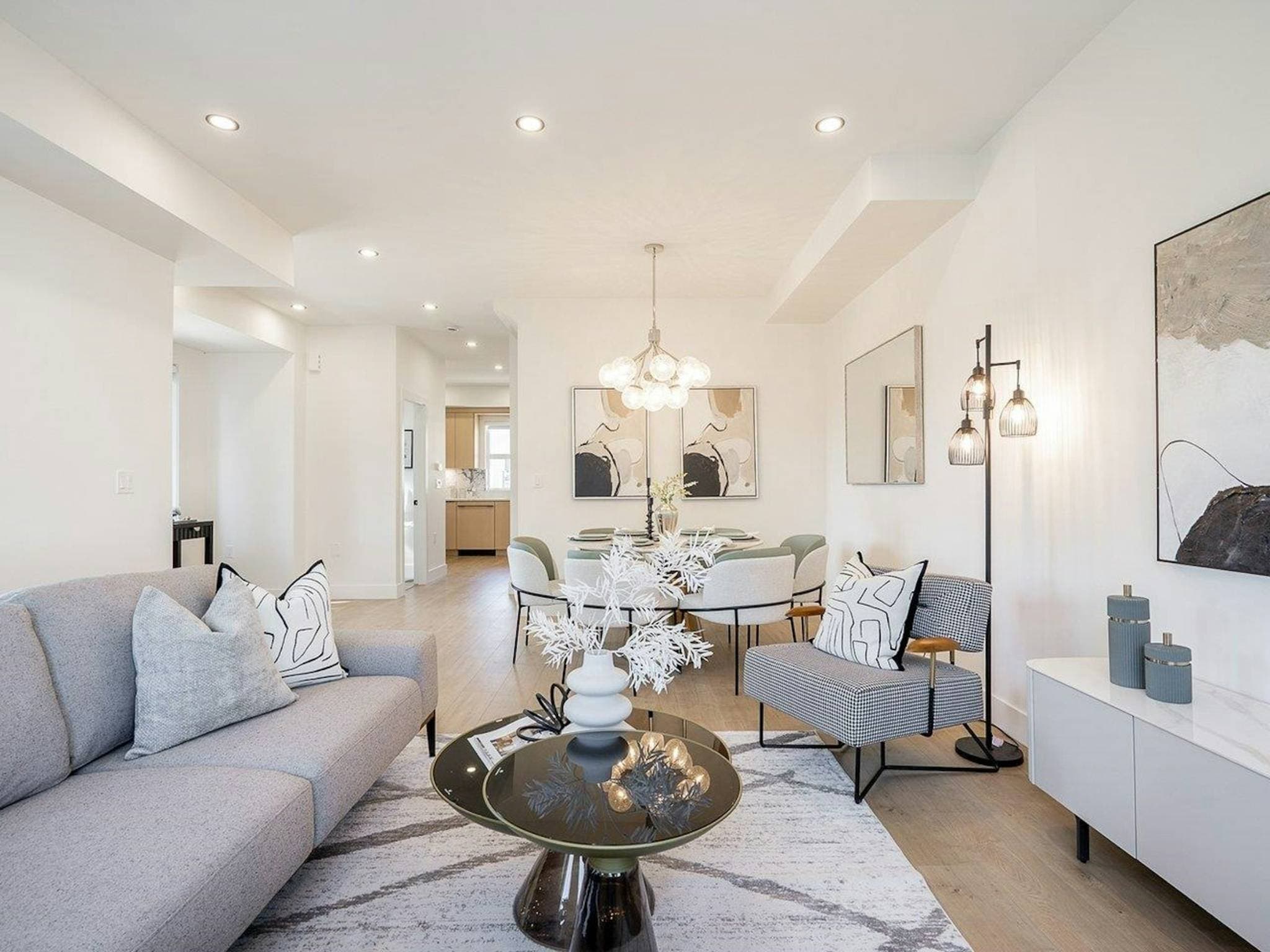Hyde Park – A bright, open-concept living and dining area featuring light wood flooring, white walls, and recessed ceiling lights. In the foreground, a grey sofa and patterned armchair surround a glass coffee table, leading past a contemporary dining set beneath a globe chandelier toward an adjacent hallway and kitchen.