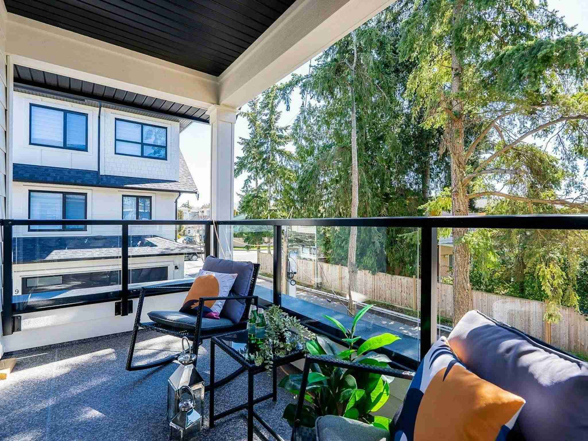 Hyde Park – A covered residential balcony or deck features modern black-framed armchairs with plush cushions, a small glass side table, potted plants, and glass railing panels. Through the railing, the view overlooks a paved street, wooden fences, tall evergreen trees, and adjacent white-sided townhomes in daylight.