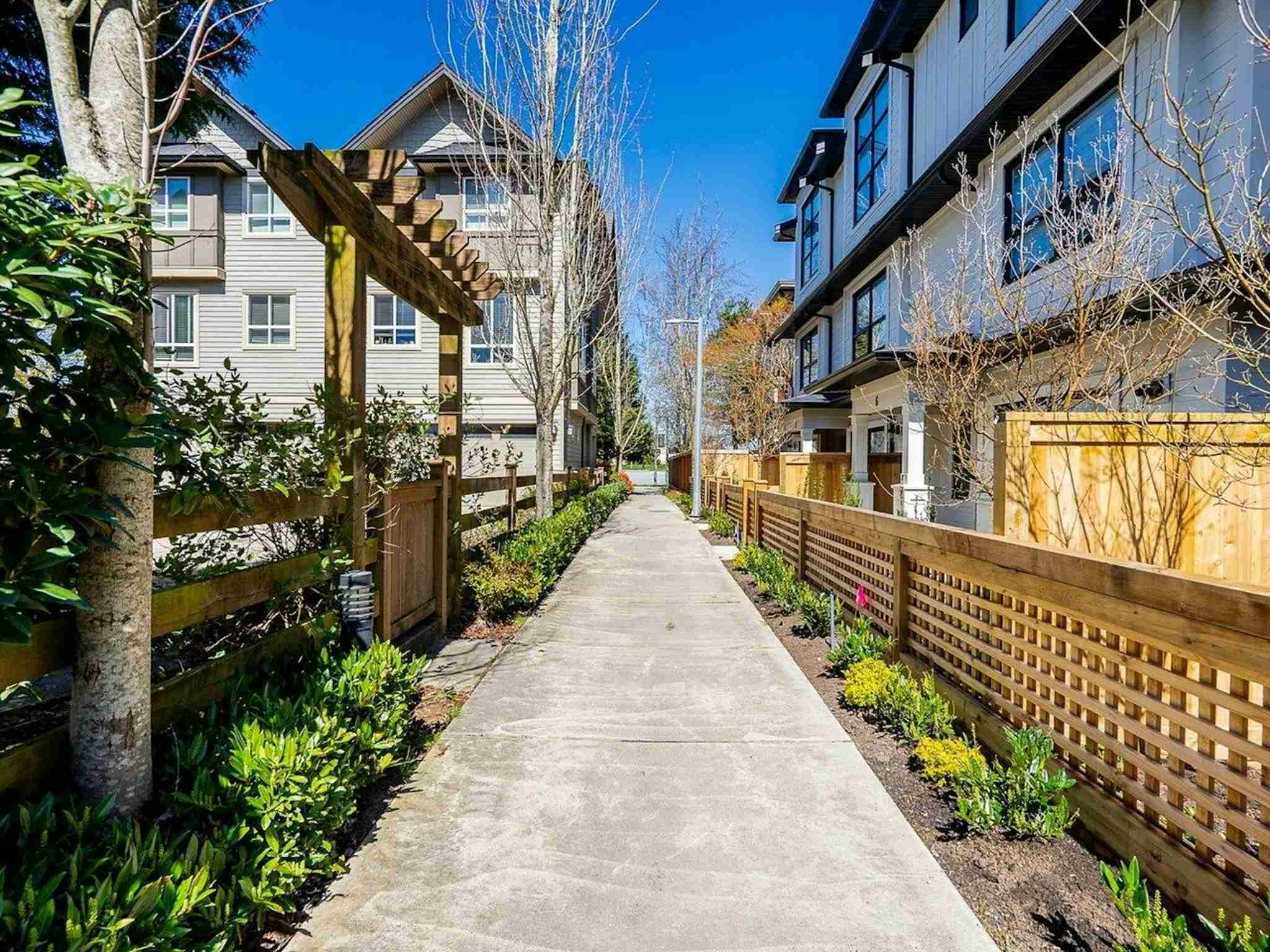 Hyde Park – A concrete pedestrian pathway runs between multi-story modern townhome complexes under a bright blue, cloudless sky. The path is bordered by wooden trellis fencing, garden beds with low shrubs, and a rustic wooden arbor gate on the left side.