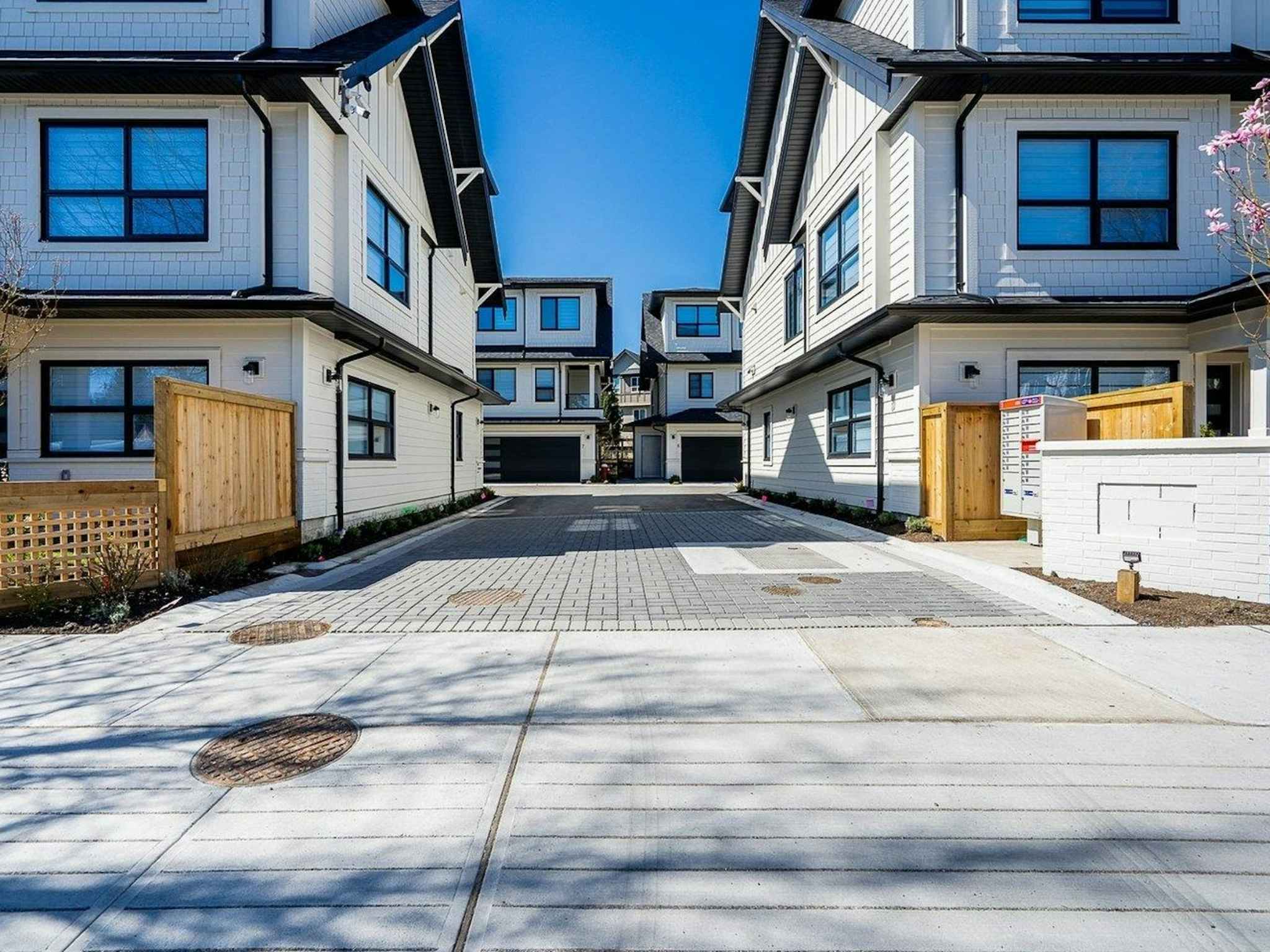 Hyde Park – A paved interior laneway flanked by modern farmhouse-style townhomes with white siding, black trim, and dark window frames under a clear blue sky. Wood privacy fencing, brick accents, and community mailboxes frame the entrance to the drive aisle.